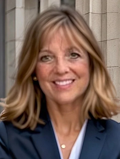 A smiling woman with shoulder-length blonde hair, wearing a navy blazer and small earrings, standing outside in front of a stone building.
