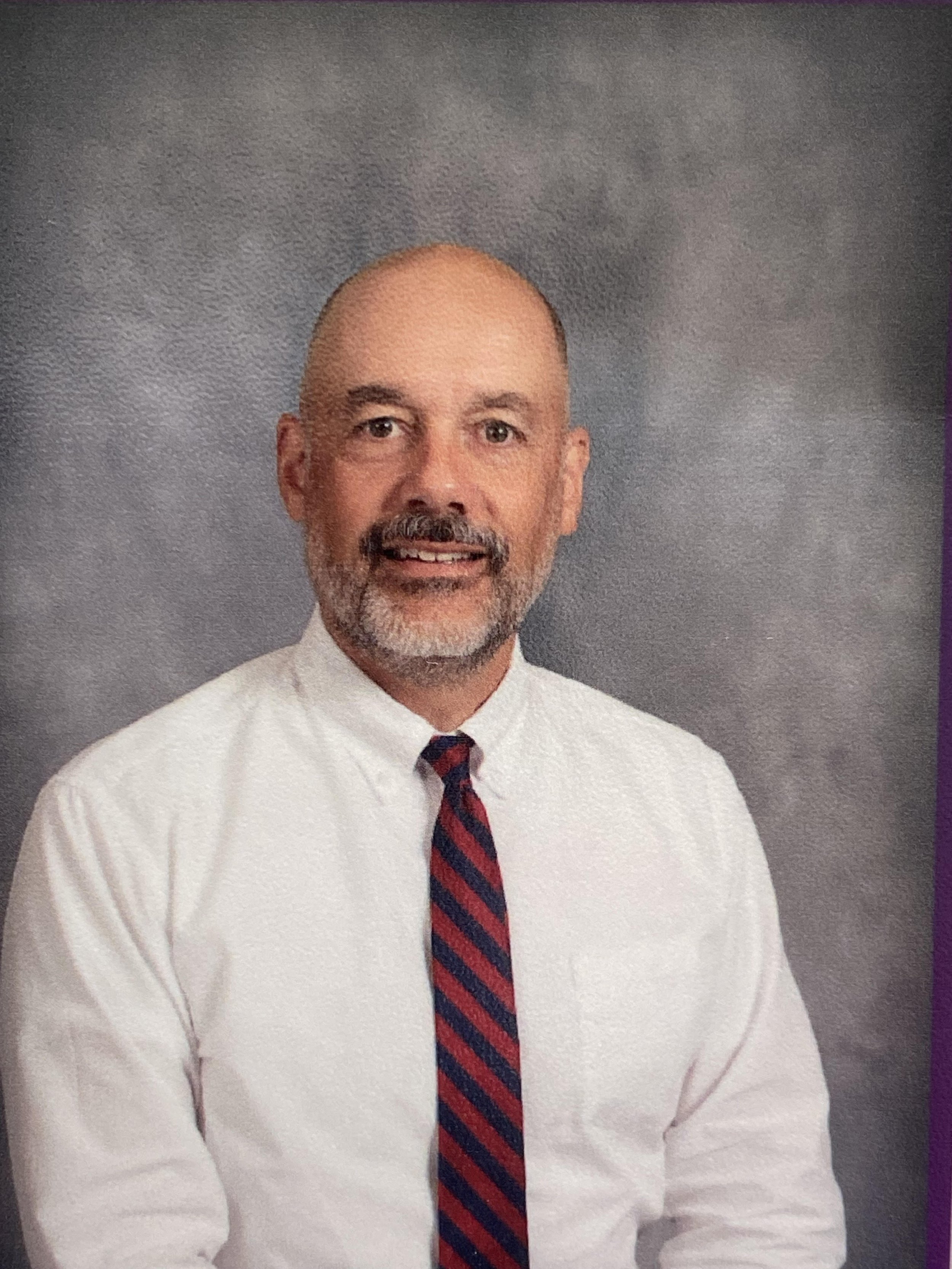 Professional headshot of a middle-aged man with a bald head, gray beard, wearing a white dress shirt and a red and navy striped tie, against a gray background.