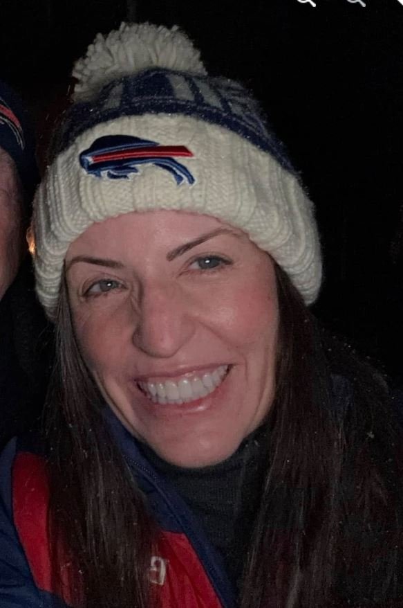 Close-up of a woman smiling outdoors at night, wearing a white knit beanie with a pom-pom and a sports team logo, by a person partially visible on the left.