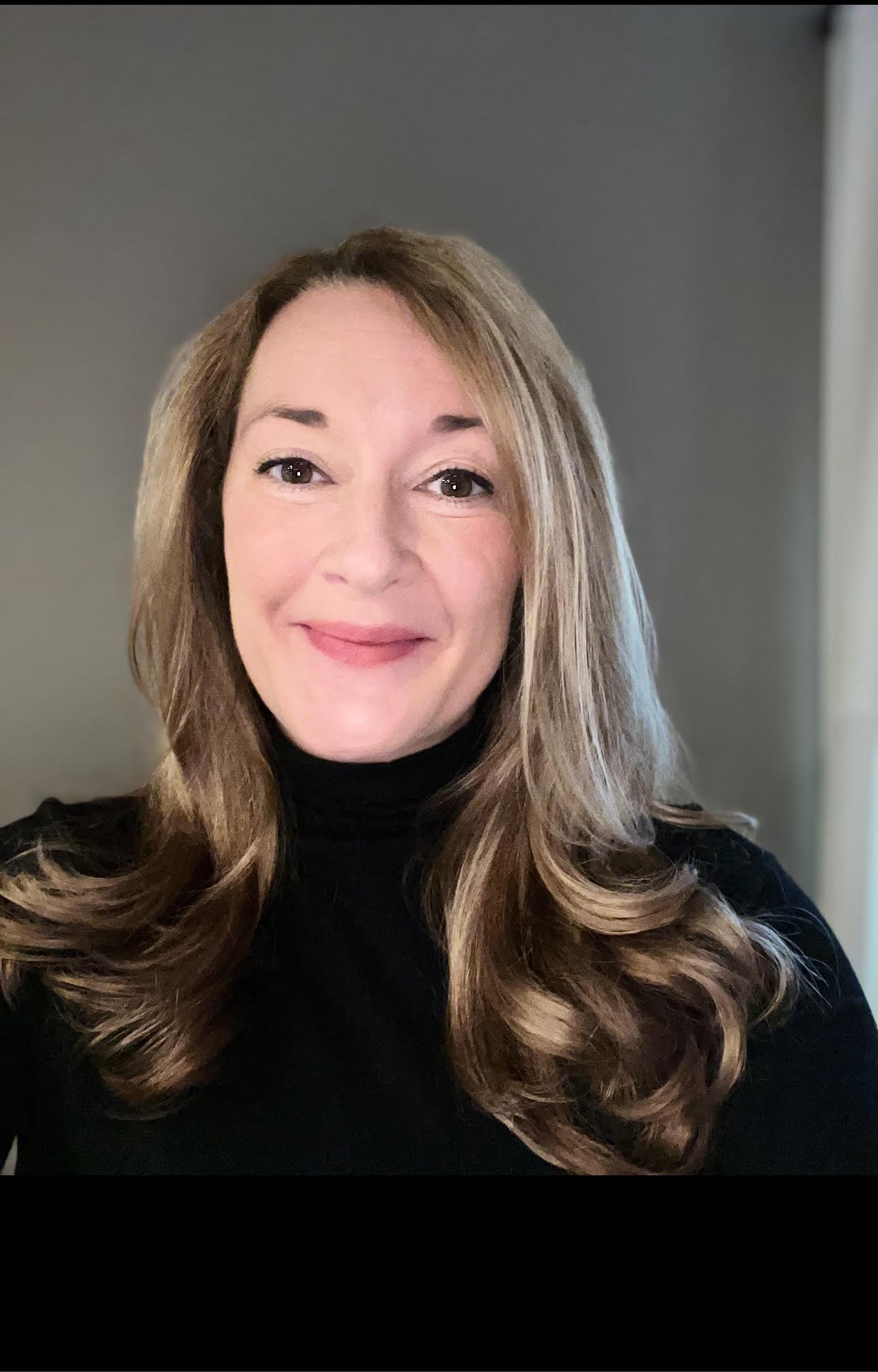 A woman with long wavy light brown hair, wearing a black turtleneck, smiling in front of a gray background.