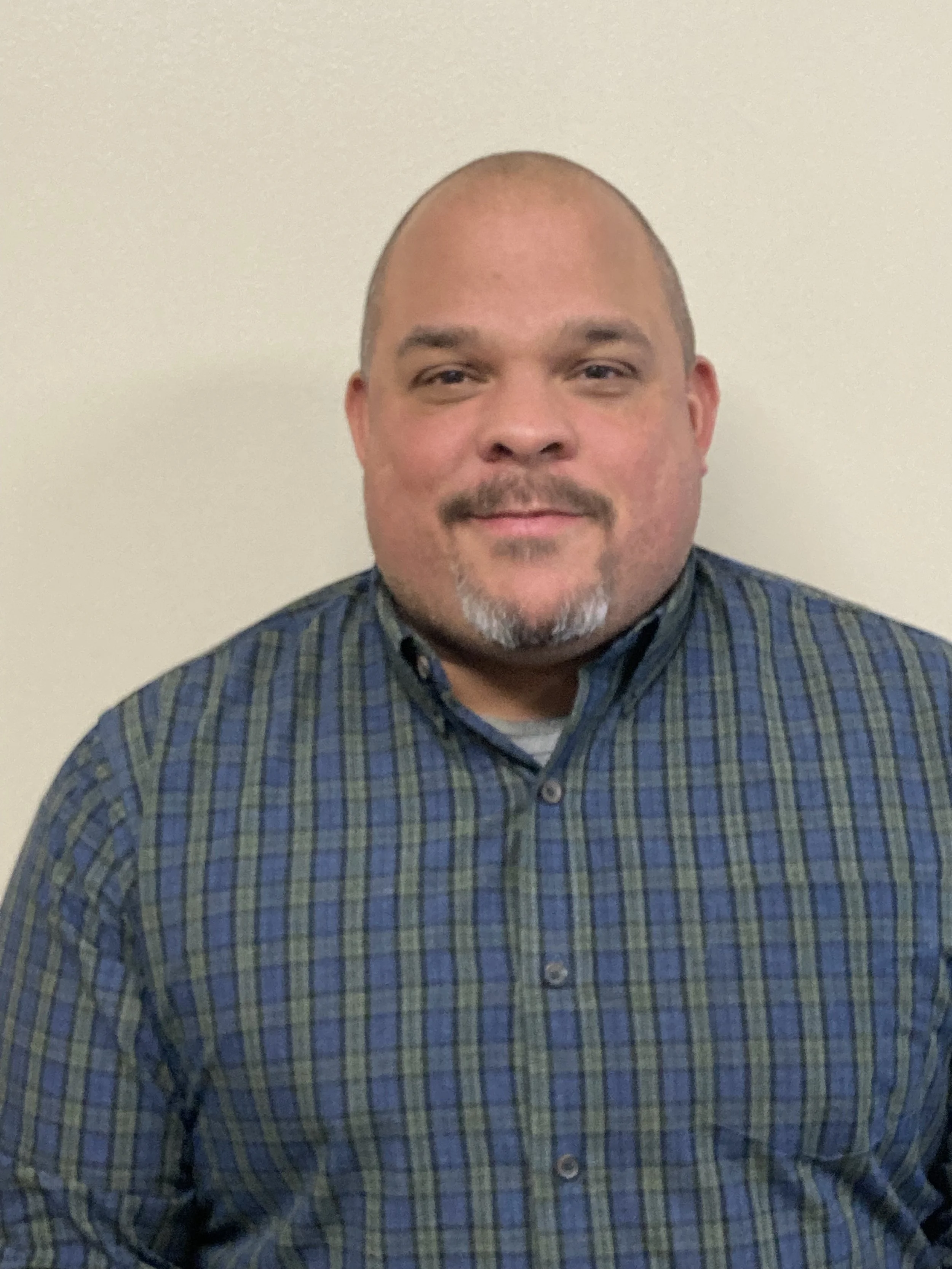A man with a beard and mustache, bald head, wearing a blue plaid button-up shirt standing against a plain white wall.