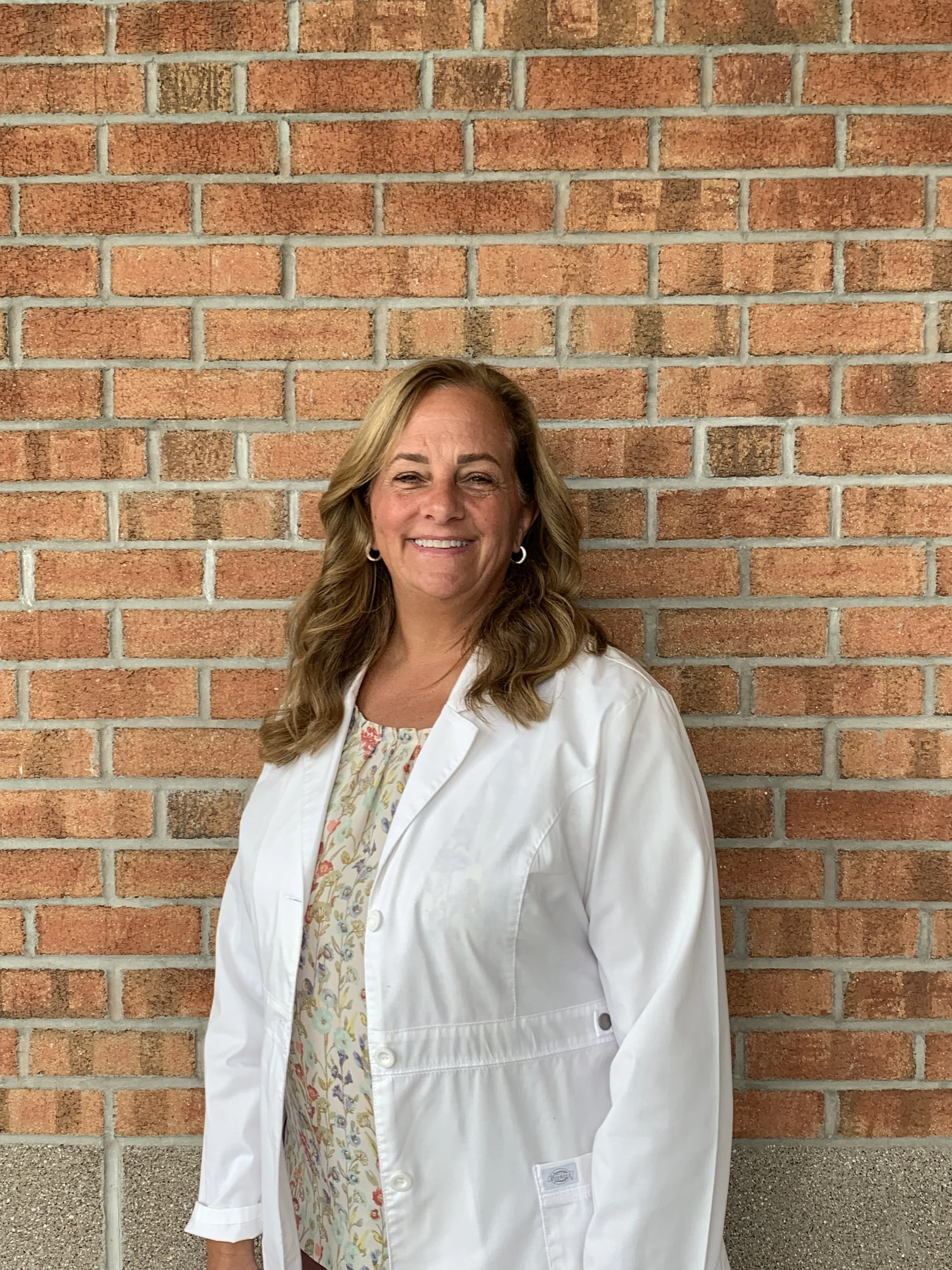 A woman in a white lab coat standing in front of a brick wall, smiling at the camera.