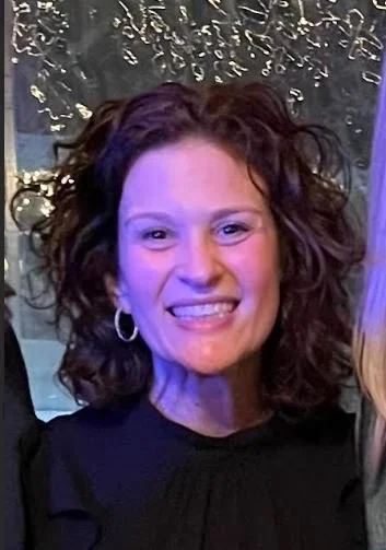 A woman with curly brown hair smiling at the camera, wearing hoop earrings and a dark top.
