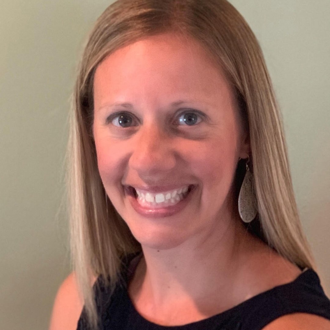 A woman with long blonde hair smiling, wearing a black sleeveless top and large silver earrings, standing against a plain background.
