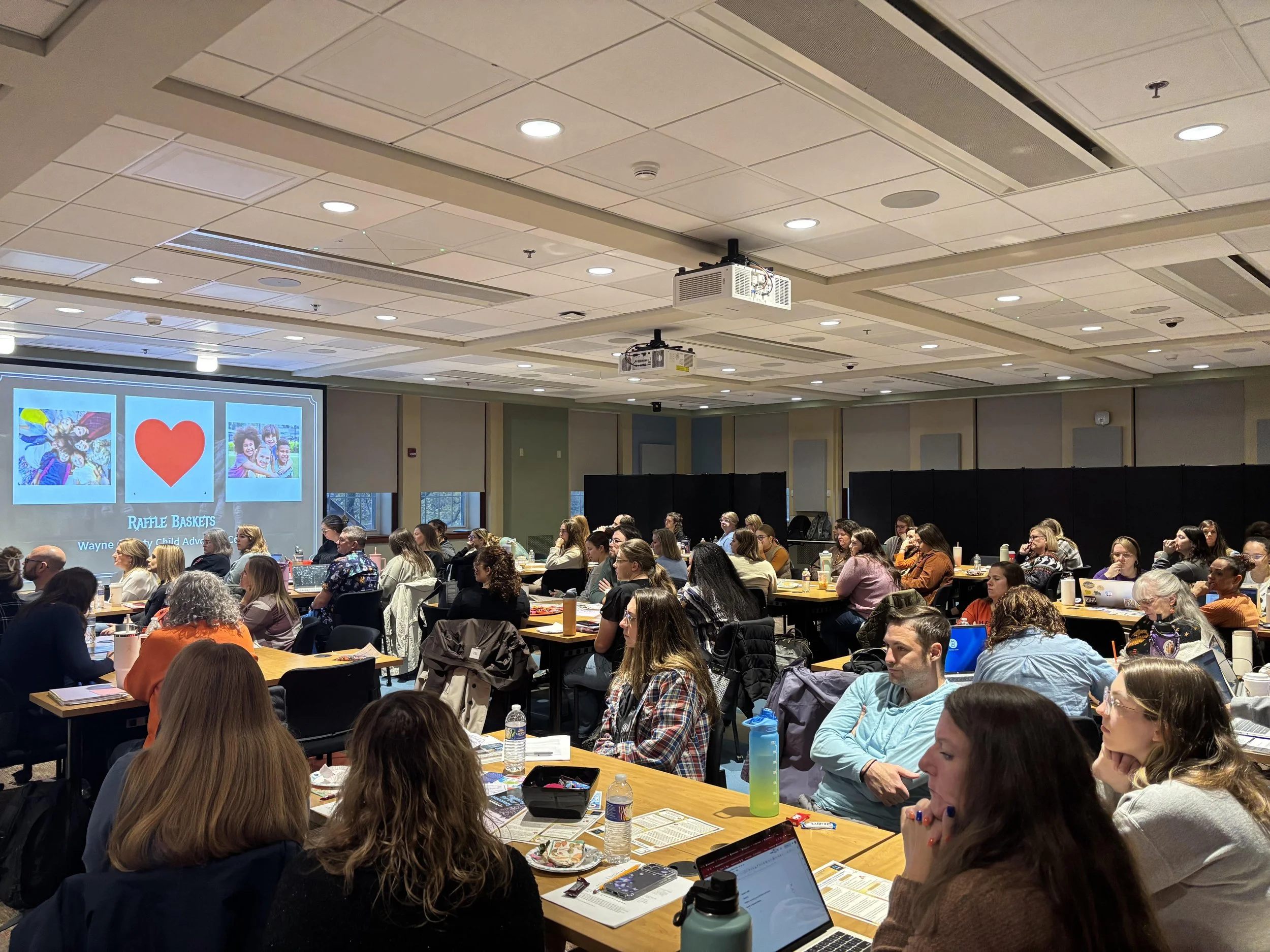 A conference room filled with attendees seated at tables, facing a presentation screen displaying images of children and a red heart with the text 'Raffle Baskets' and 'Wayne County Child Advocacy Center'.