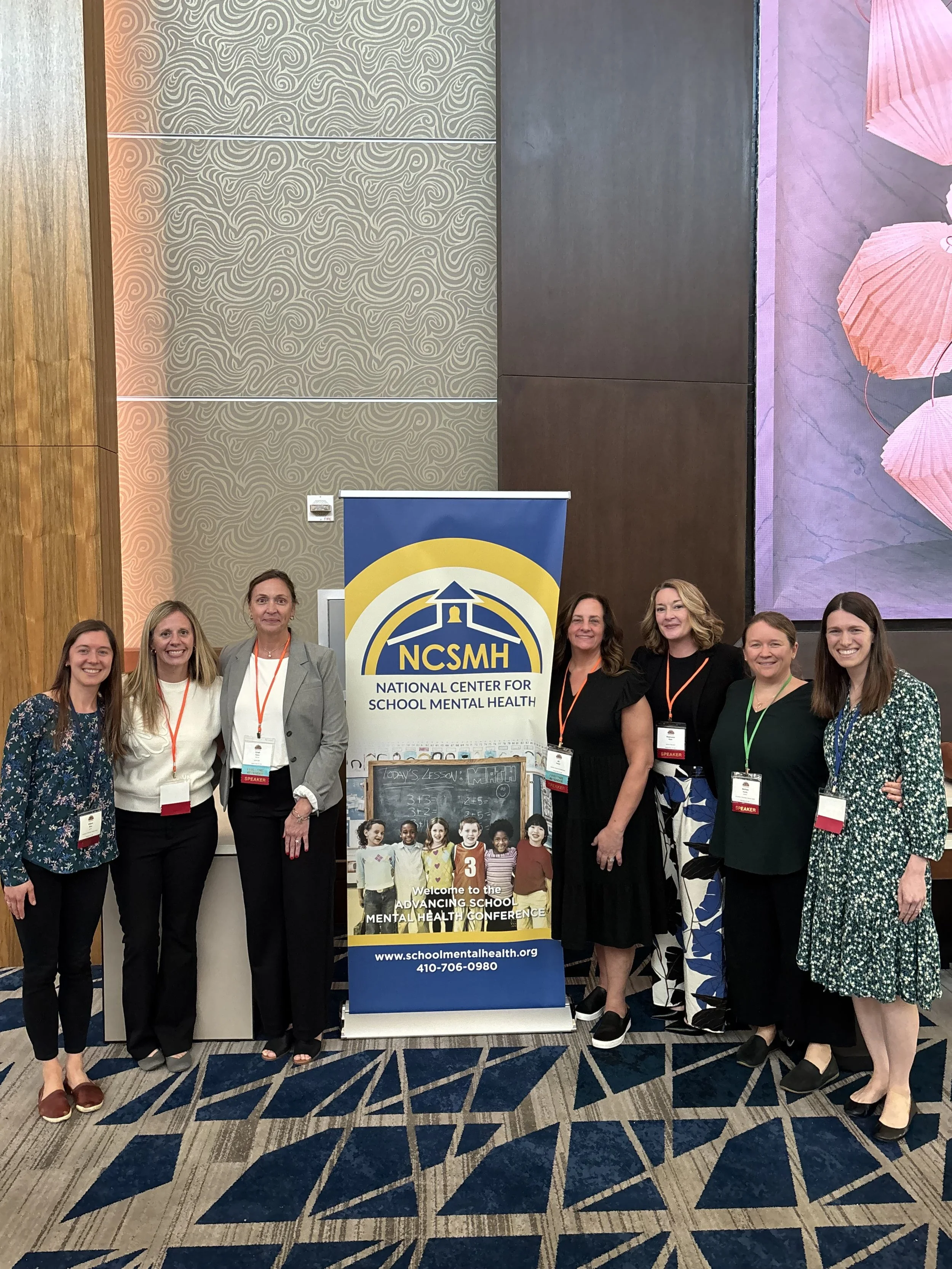 Group of eight women standing beside a banner for the National Center for School Mental Health at a conference, posing indoors on a patterned carpet.