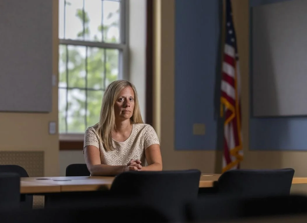 A woman with long blonde hair sitting alone at a table in a room with blue and yellow walls, large window with natural light, American flag in the background.