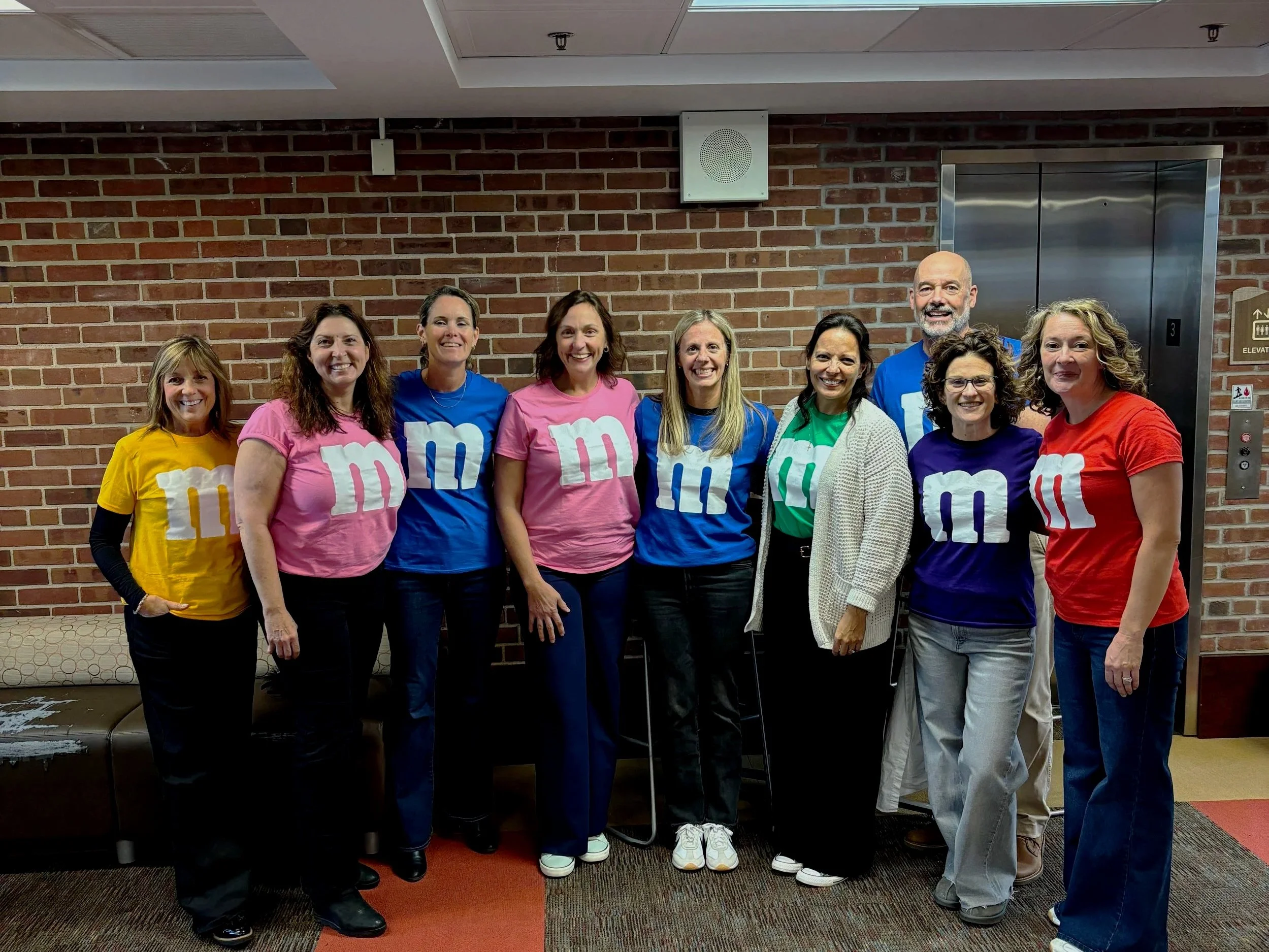 Group of nine women and one man standing in front of a brick wall, wearing colorful M&M's themed T-shirts, smiling.