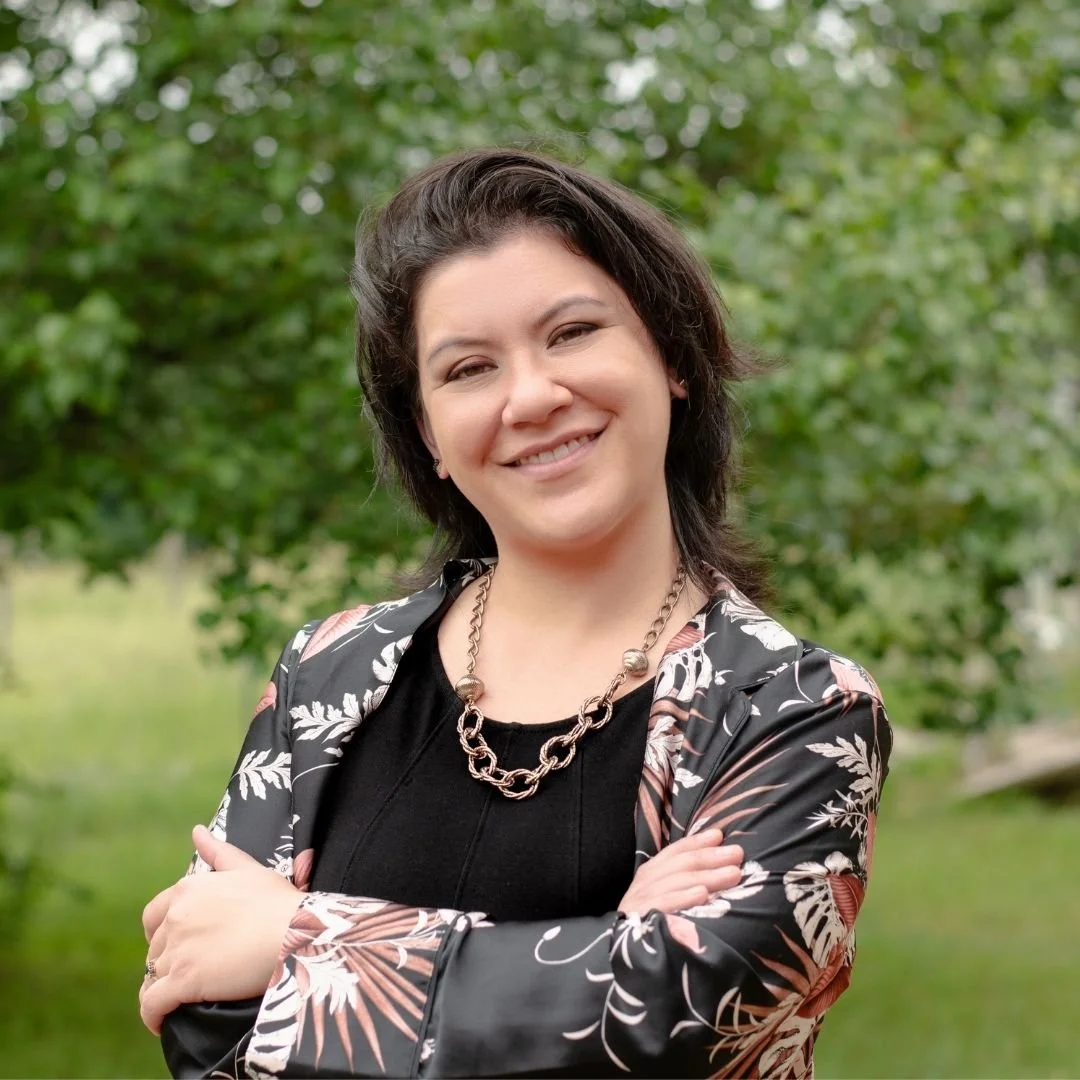 Gabby Hanson smiling outdoors, wearing a black top, a patterned blazer, and a chain necklace, with green trees in the background.