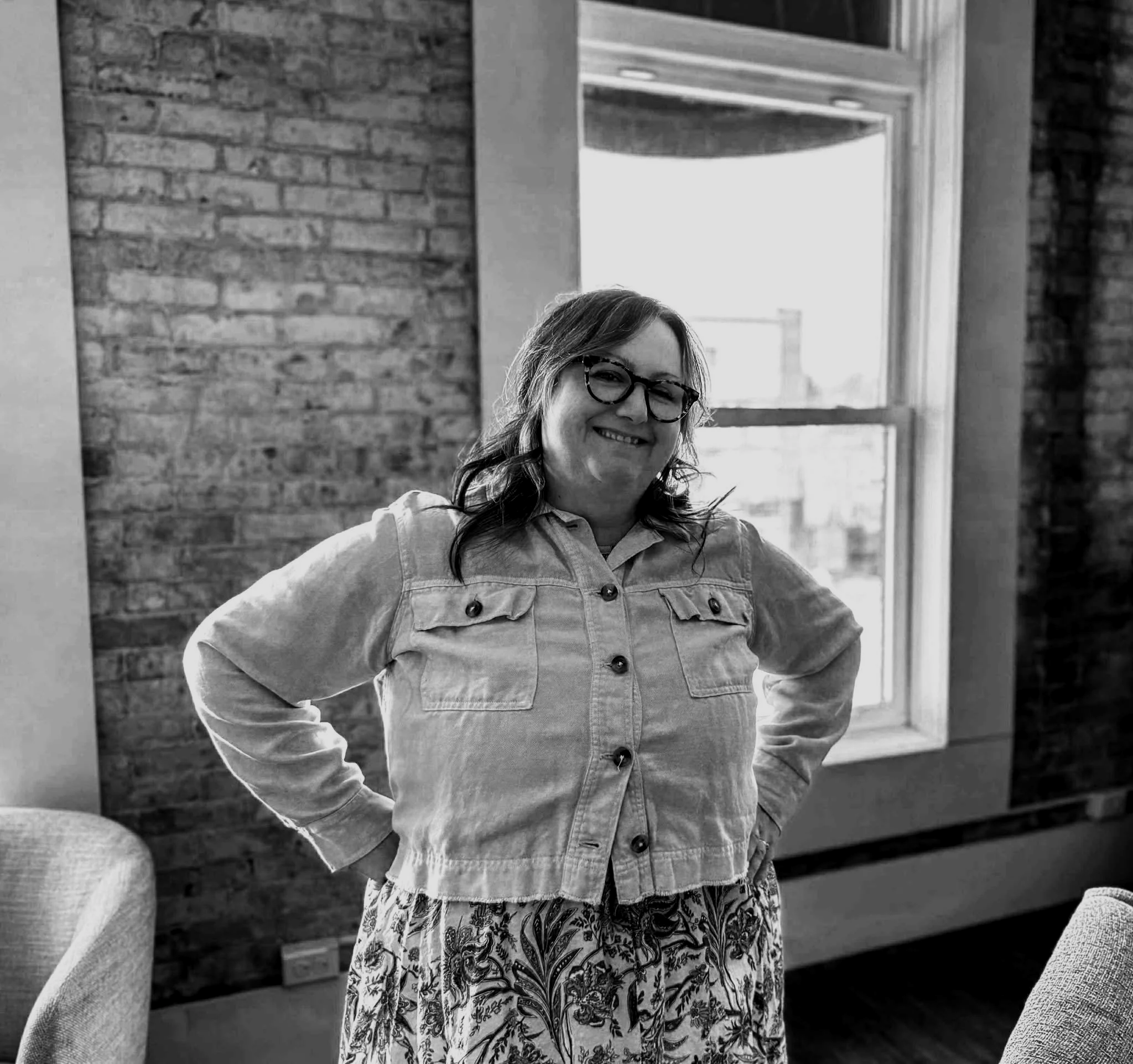 Dani Renwick smiling, wearing a denim jacket and floral skirt, standing indoors near a window with her hands on her hips.