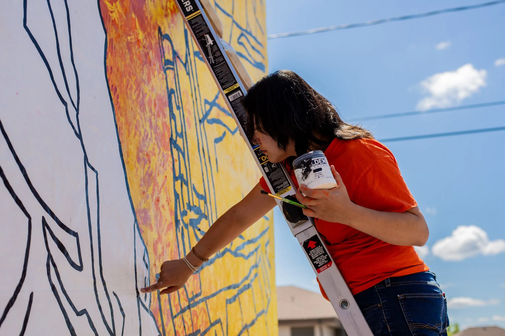 A woman in an orange shirt and jeans is painting a colorful mural on a wall outdoors. She is holding a container of white paint and a paintbrush, with a ladder propped against the wall. The mural features pathways and trees in bright colors. The sky is blue with some clouds.