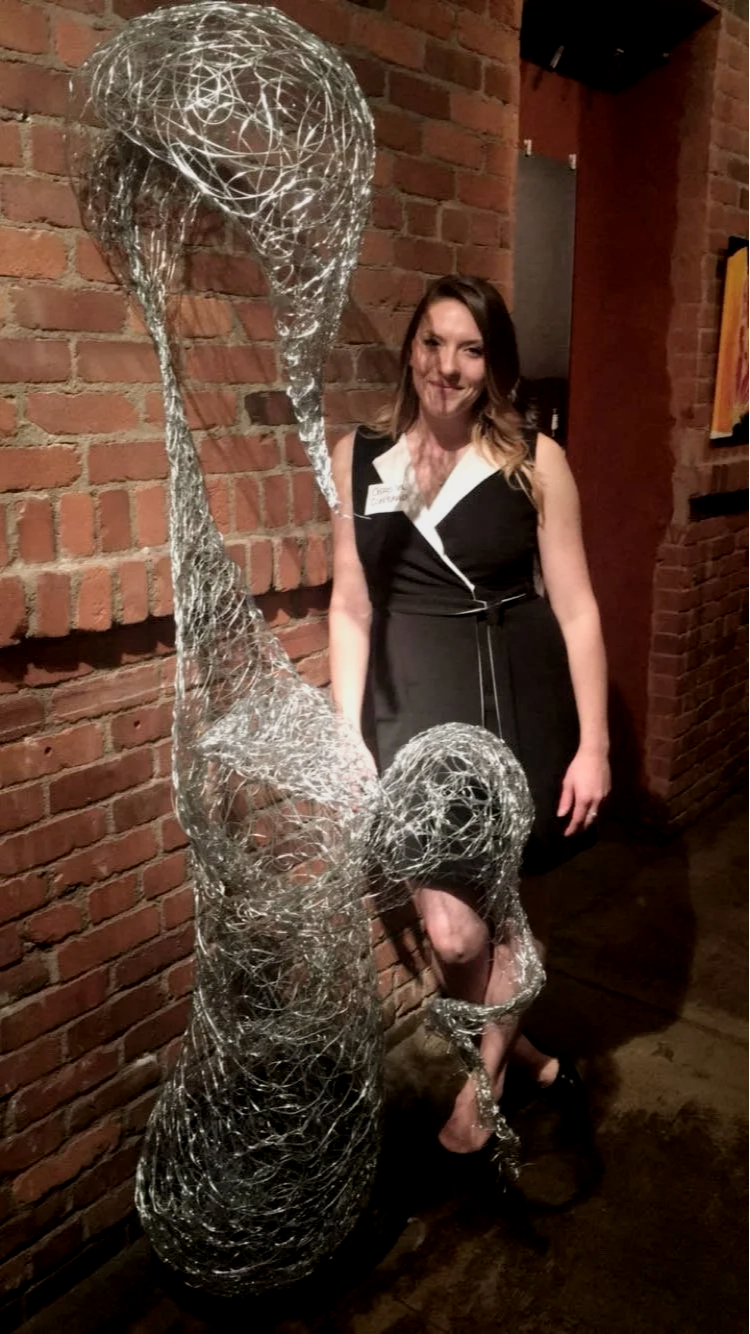Christen Contario in a black dress standing next to a large abstract wire sculpture resembling a flower against a brick wall.