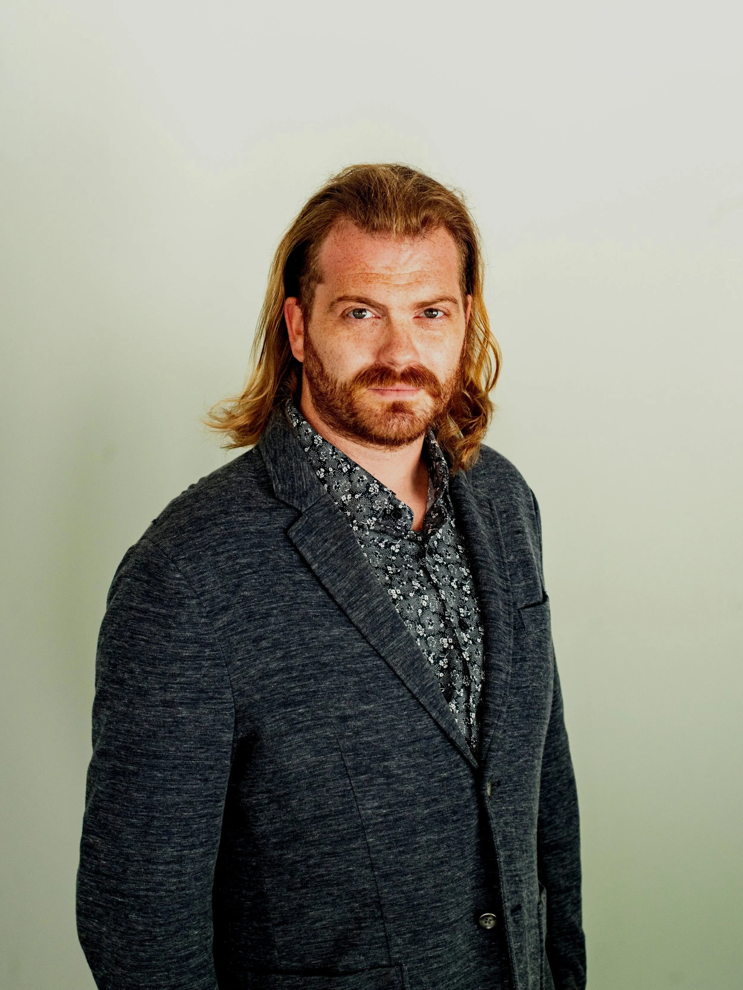 Heath Urbaniak wearing a dark blazer over a patterned shirt, standing against a plain, light-colored background.