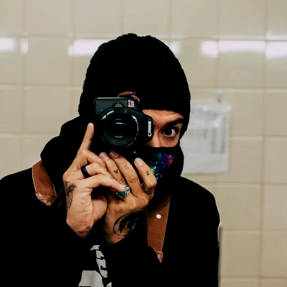 Anthony SlaughterHawk wearing a black beanie and black face mask with a galaxy print, taking a selfie with a Canon camera in a room with beige tiled walls.