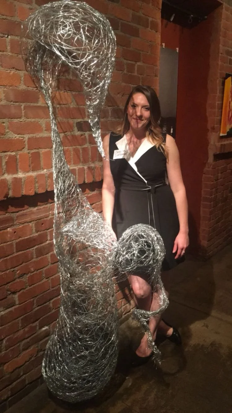 Christen Contario standing next to an abstract sculpture made of silver wire against a brick wall.