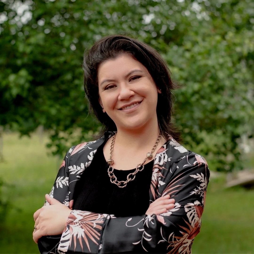 Gabby Hanson smiling with arms crossed, outdoors with green trees in the background.
