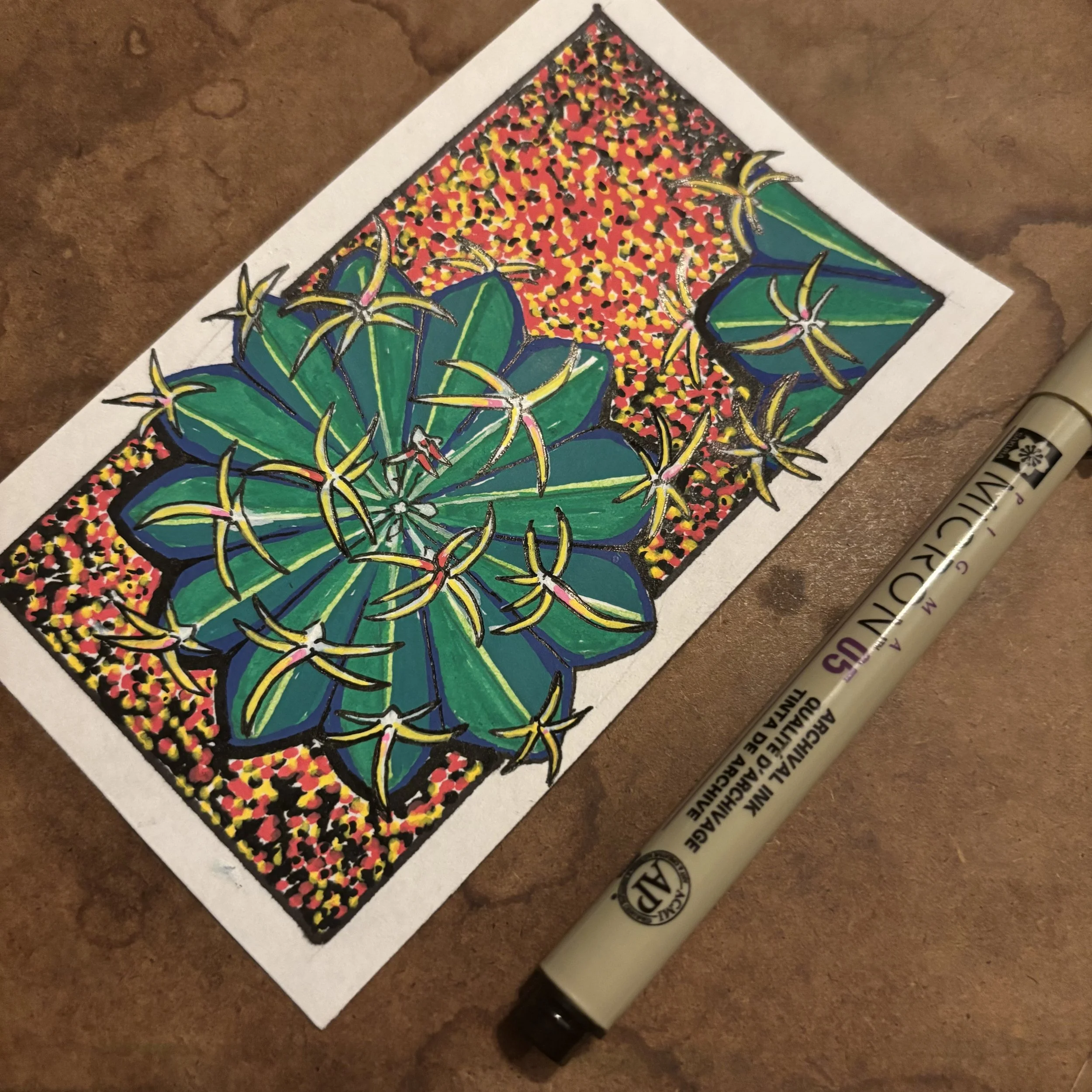 A colorful drawing of two large green succulents with yellow-tipped leaves, surrounded by a red, yellow, and black dotted background, on a piece of paper resting on a brown surface, with a beige black marker lying beside it.