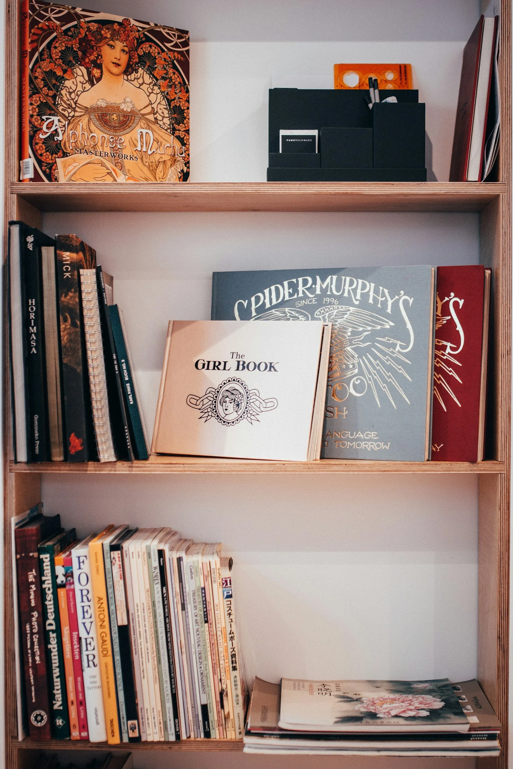 A wooden bookshelf filled with various books, magazines, and decorative items. The top shelf has an art book with a colorful cover featuring an illustrated woman. The middle shelf holds several books, including 'The Girl Book' and 'Spider Murphy's,' with some arranged upright and others leaning. The bottom shelf contains more books, magazines, and possibly a newspaper or catalog.