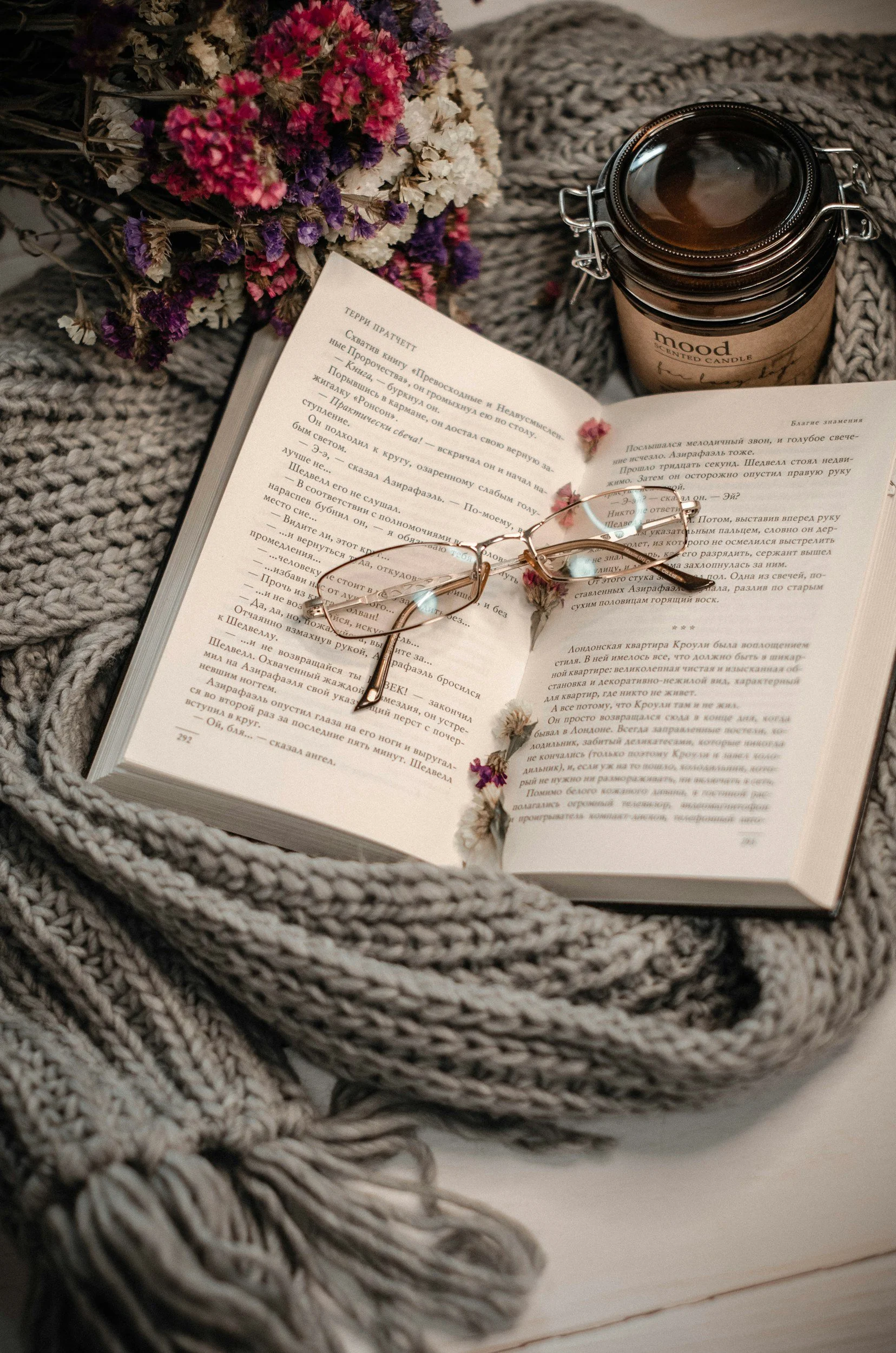 An open book with text in a foreign language, a pair of glasses resting on the pages, surrounded by a chunky knit blanket, a bouquet of colorful dried flowers, and a candle in a brown jar labeled 'mood'.