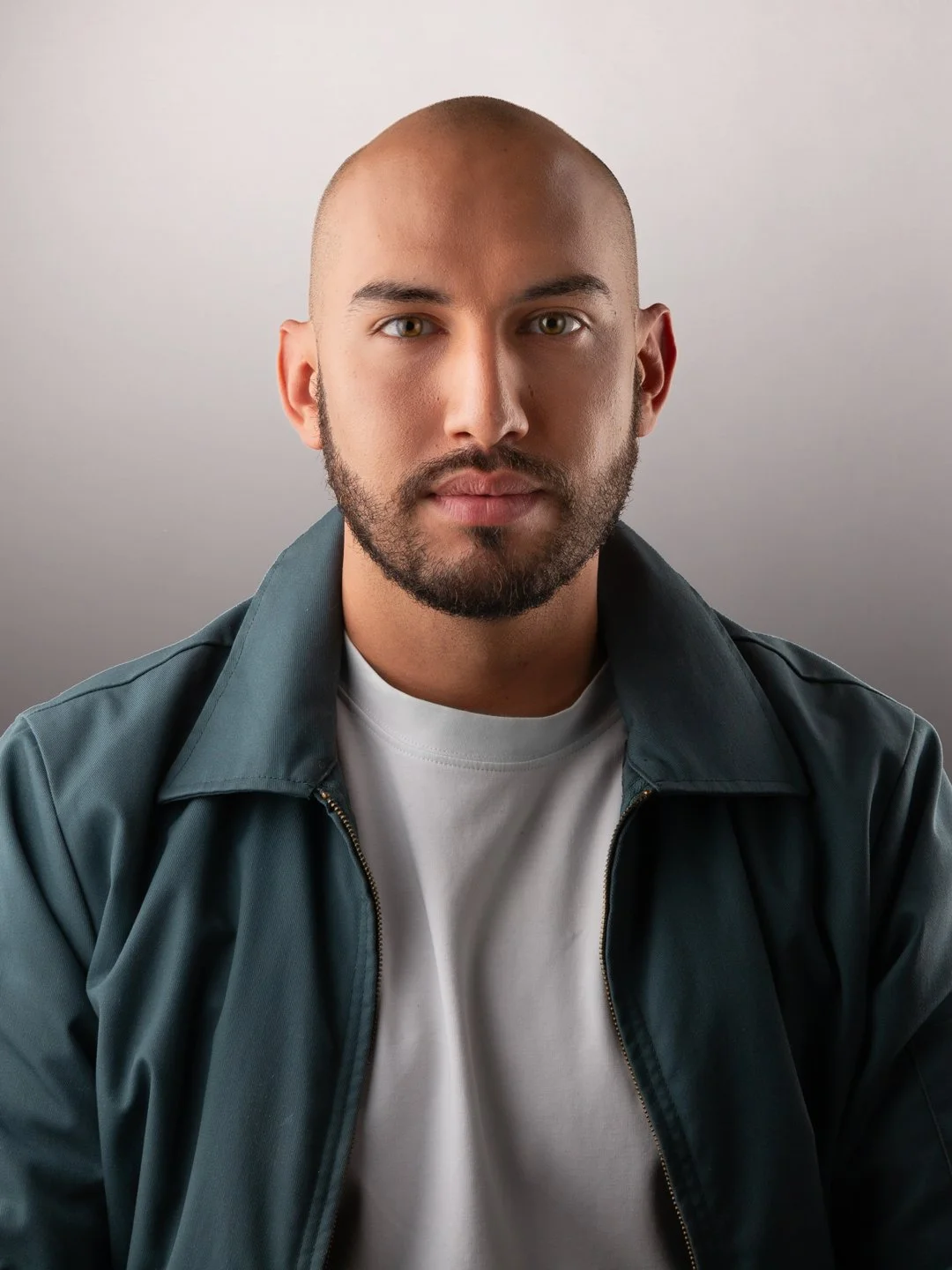 Retrato de un hombre con barba, sin cabello y con expresión neutral, vistiendo una chaqueta oscura y camiseta blanca, en un fondo gris claro.