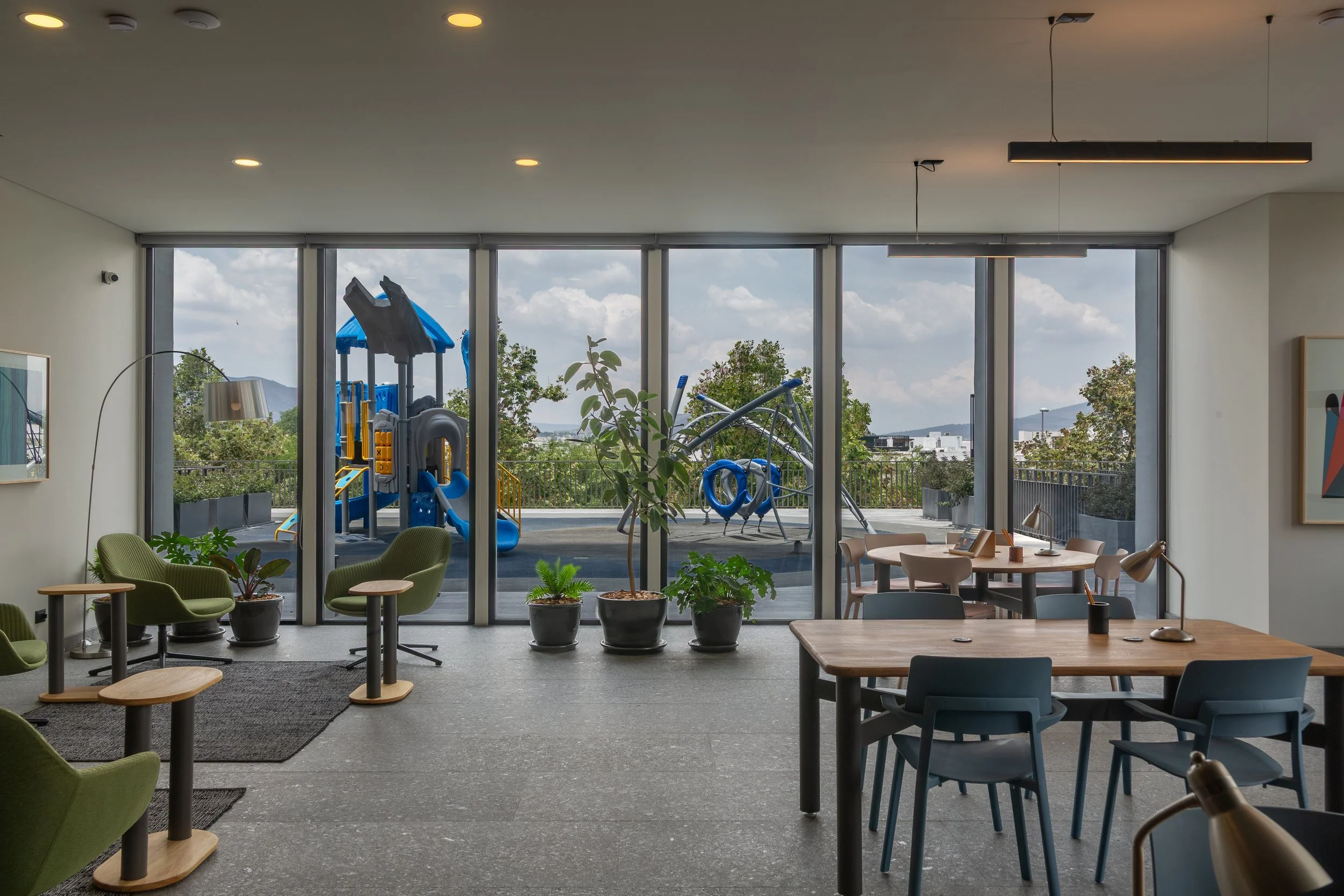Sala de estudio o trabajo con ventanas grandes que dan a un área de juegos para niños con estructura de plástico azul y andador. Tiene varias sillas y mesas, plantas decorativas y cuadros en las paredes.