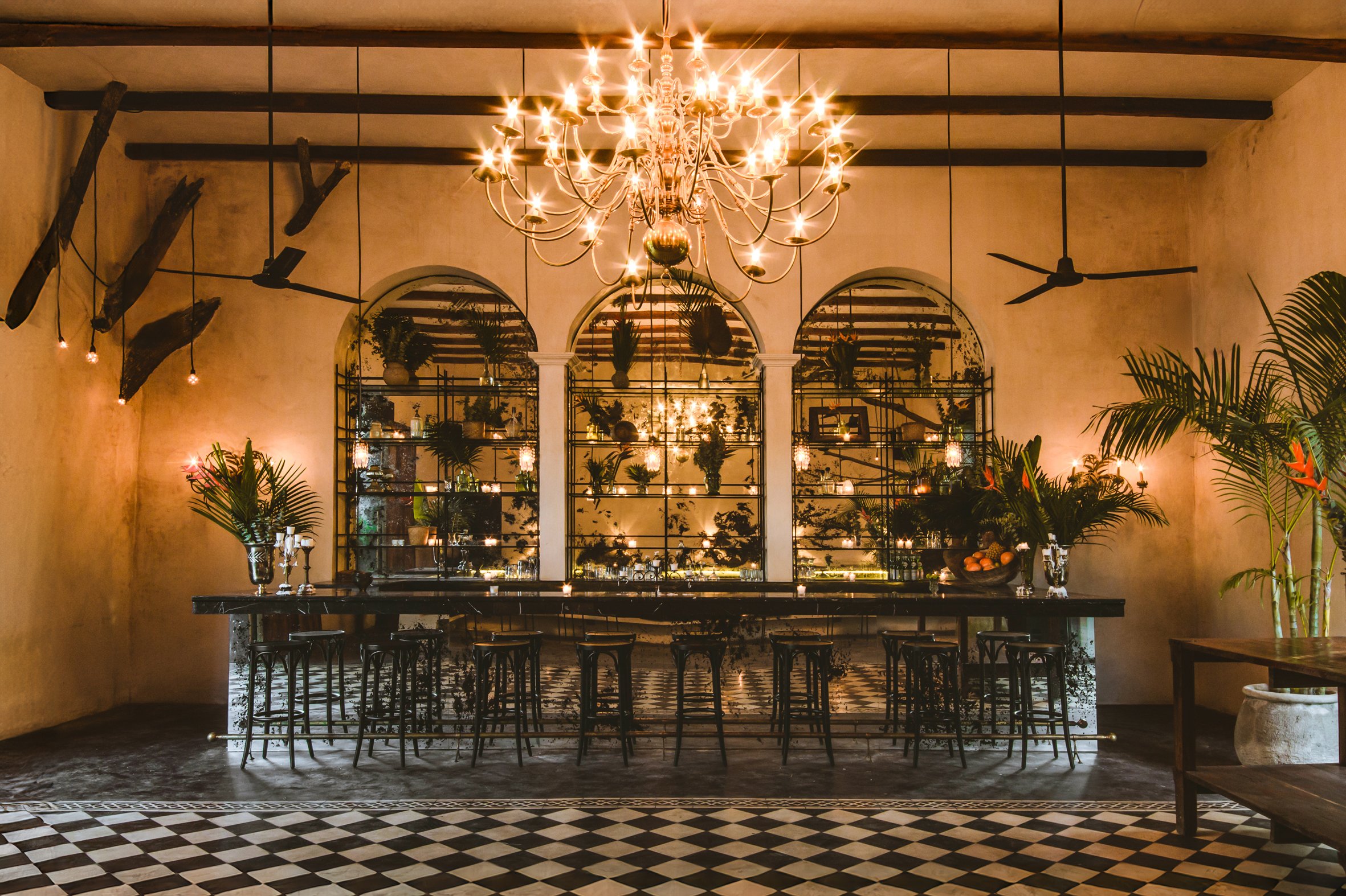 Interior elegante con lámpara de araña, espejos, estanterías con plantas y decoración luminosa, mesa larga con sillas, plantas decorativas, paredes de color beige y suelo a cuadros.