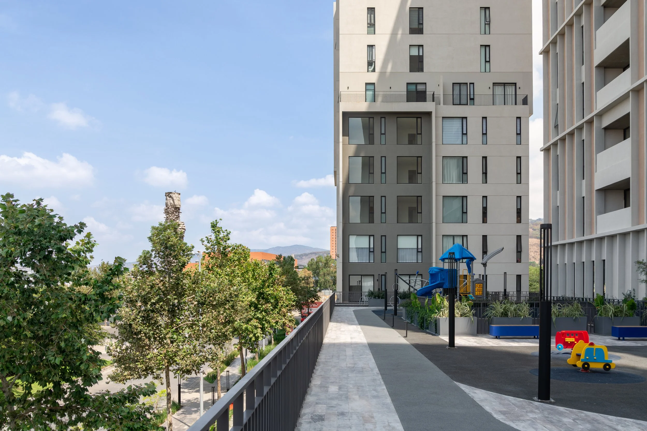 Vista de un complejo de apartamentos modernos con área de juegos infantiles, árboles y un cielo azul con nubes.