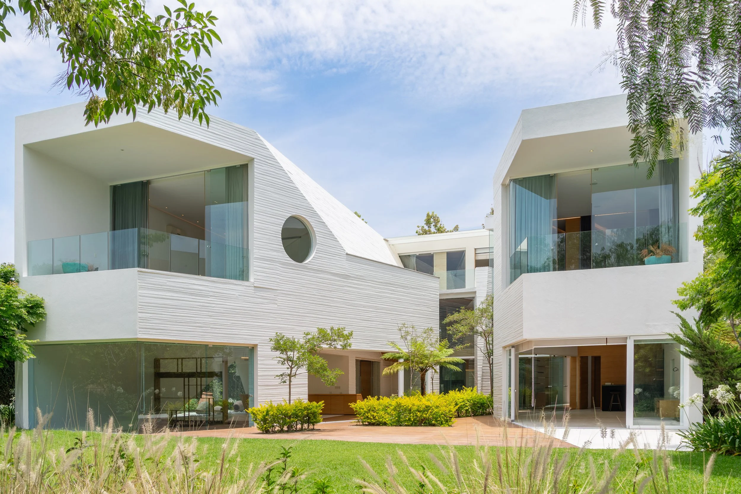 Casa moderna de construcción blanca con amplios ventanales, en medio de jardín con arbustos y árboles, bajo cielo despejado.
