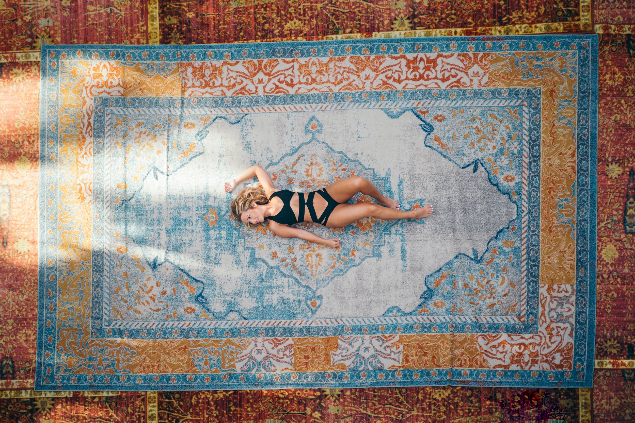 Mujer joven en bikini negro acostada en una alfombra decorativa de colores vivos y patrones elaborados, en una habitación con alfombras distintas, vista desde arriba.