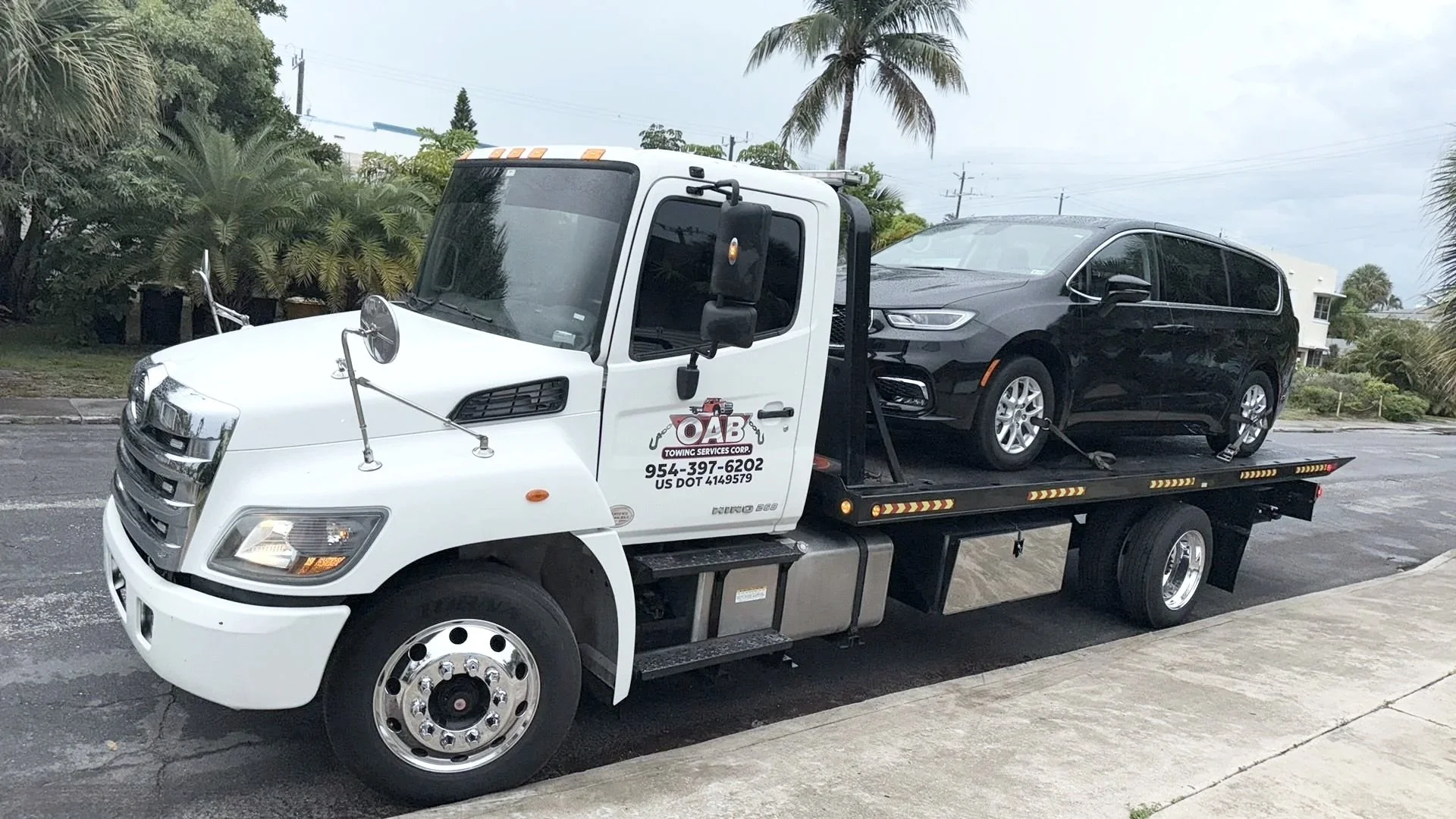 A flatbed tow truck carrying a black minivan on a wet road with palm trees and houses in the background.