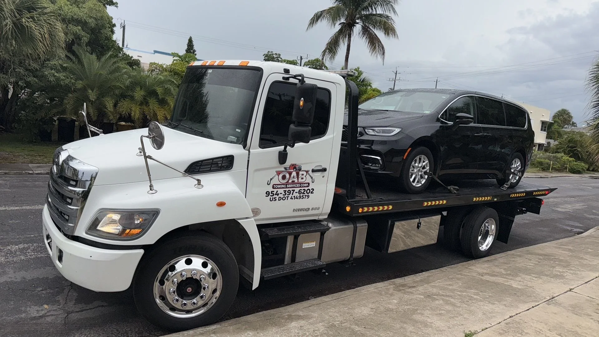 A tow truck carrying a black minivan, parked on a street with palm trees and cloudy sky in the background.