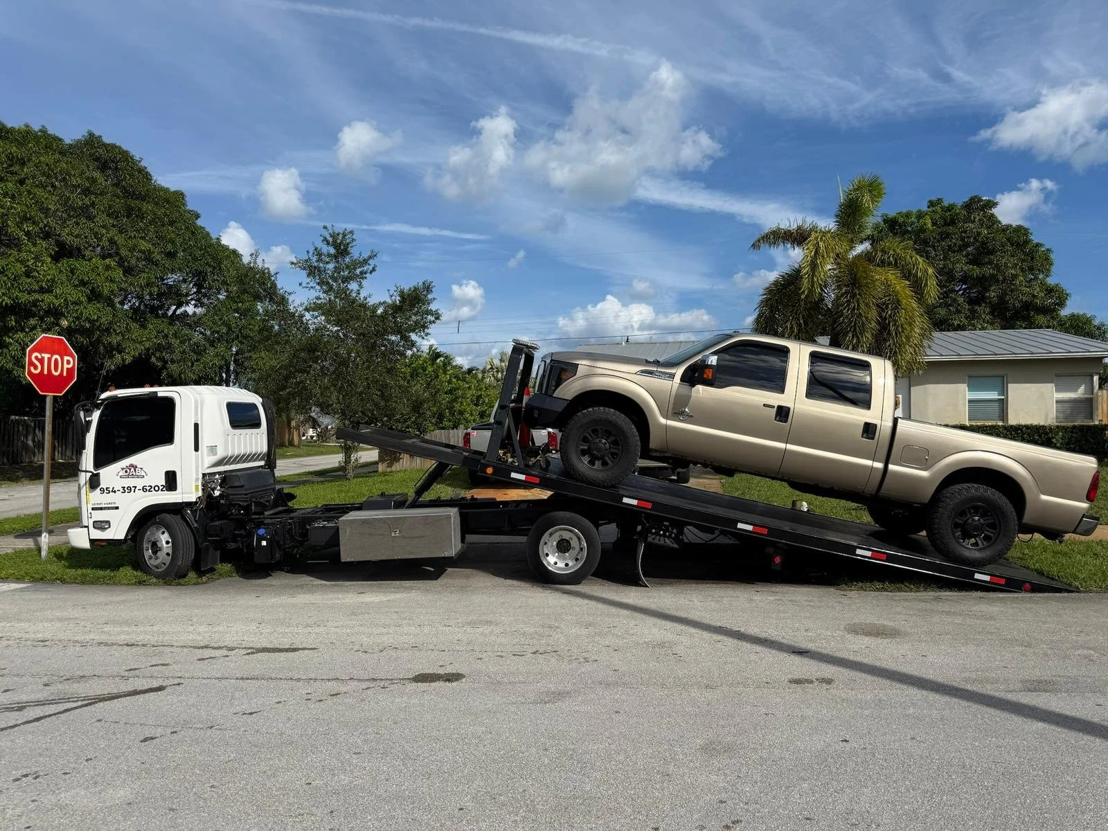 A tow truck transporting a beige pickup truck in a residential neighborhood, with trees, a house, and a blue sky with clouds in the background.