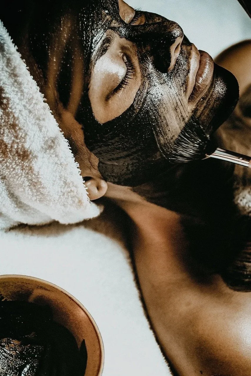 A woman with dark hair is receiving a facial treatment with a black mud mask, lying down with her eyes closed and a towel wrapped around her head, as a spa professional applies the mask.