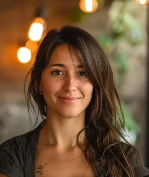 A young woman with long wavy brown hair smiling at the camera, with blurred warm lights and greenery in the background.