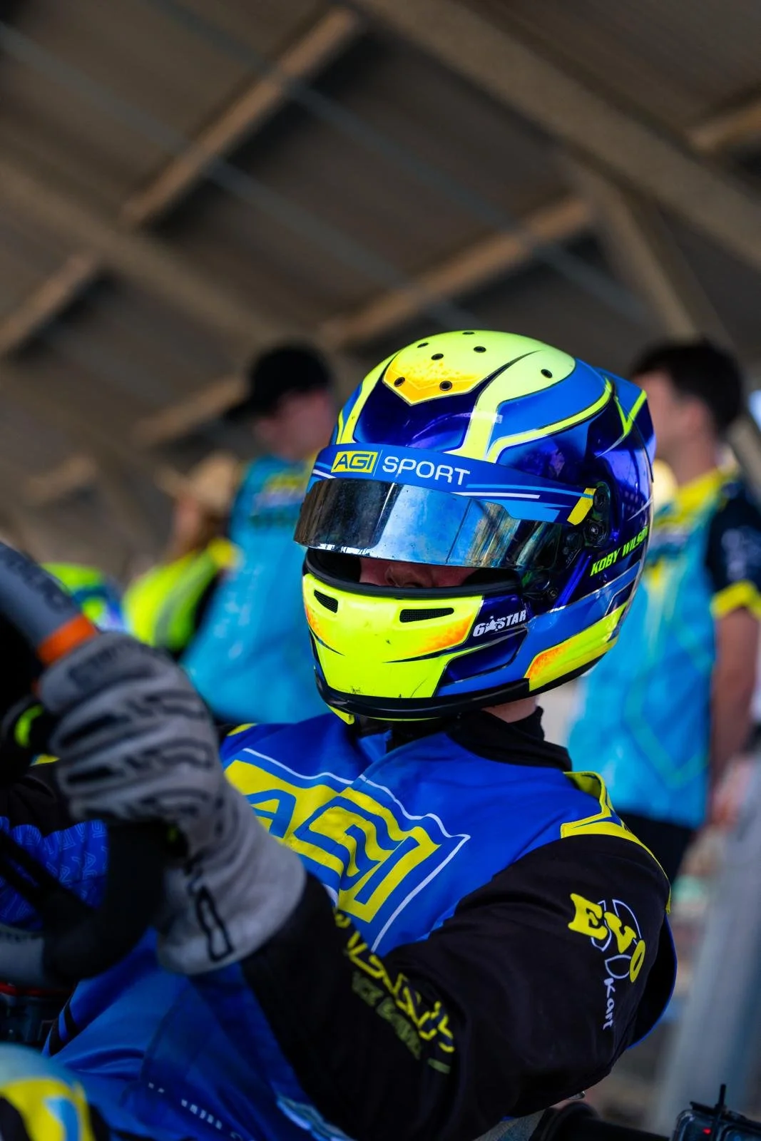 A race car driver wearing a blue and yellow helmet and racing suit, with people in the background under a wooden roof.
