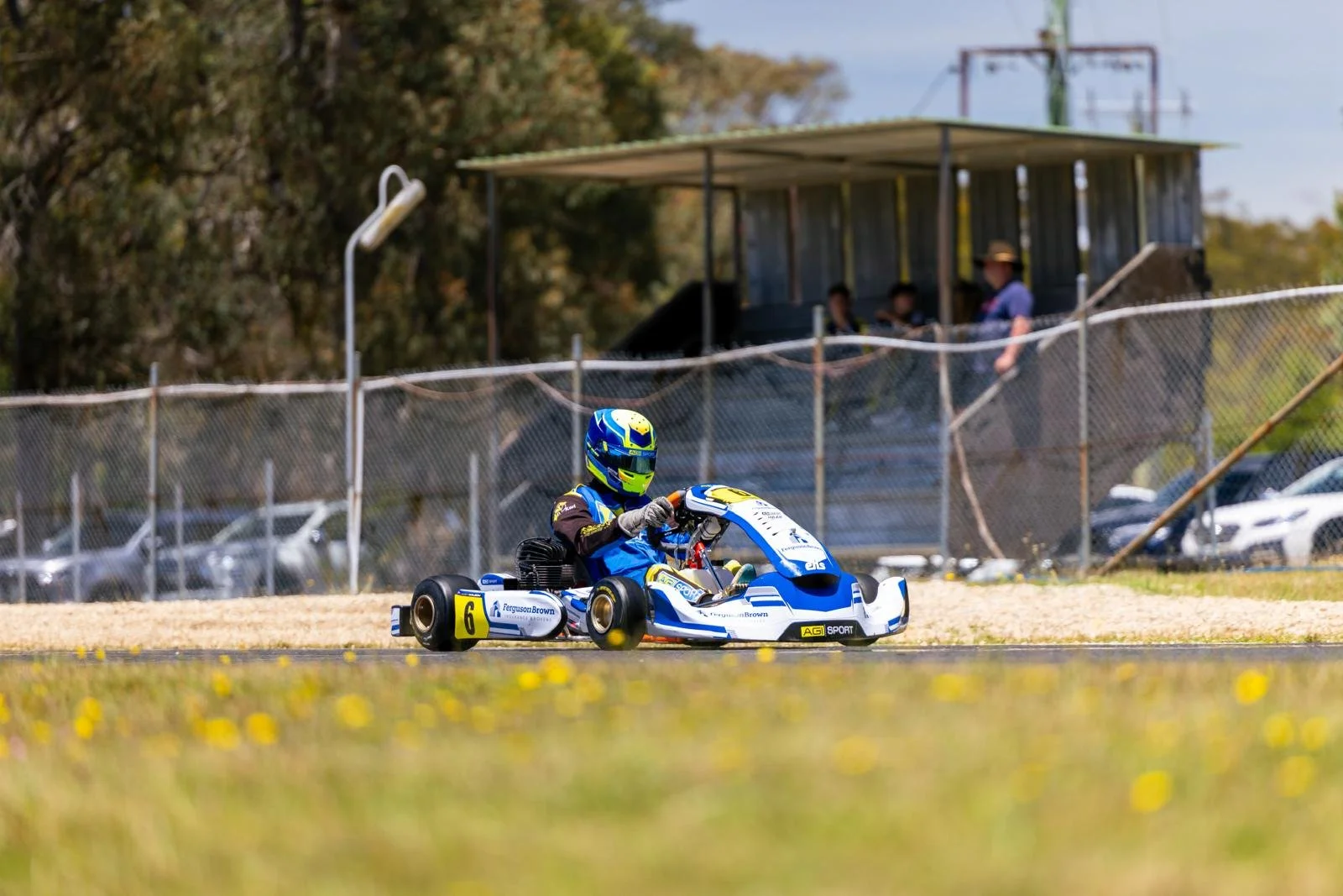 A person in a blue racing suit and helmet driving a blue and white go-kart on a race track, with a fence and spectators in the background.