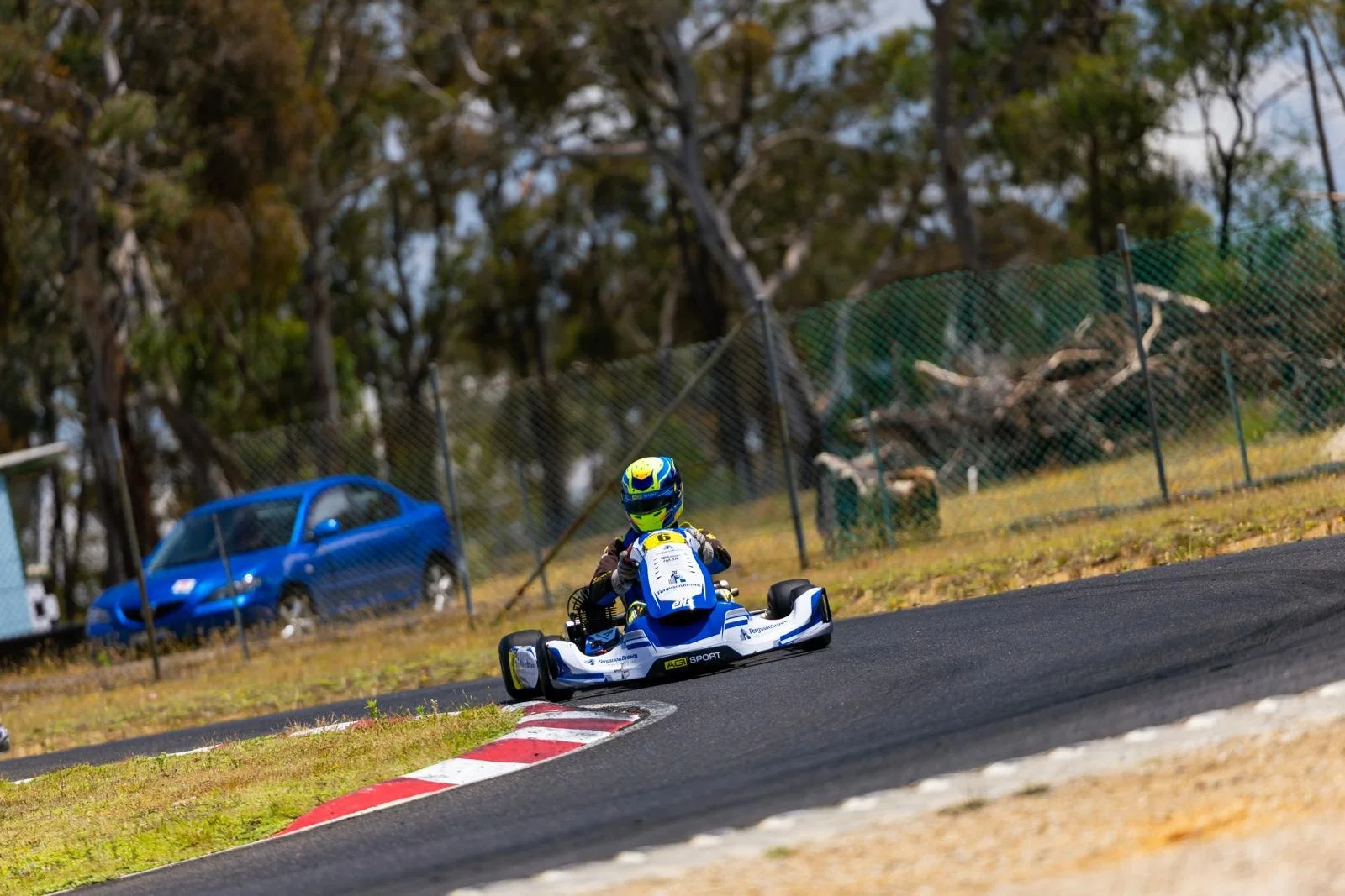 A go-kart racer wearing a blue and yellow helmet and racing suit navigates a turn on a race track, with a blue car and trees in the background.