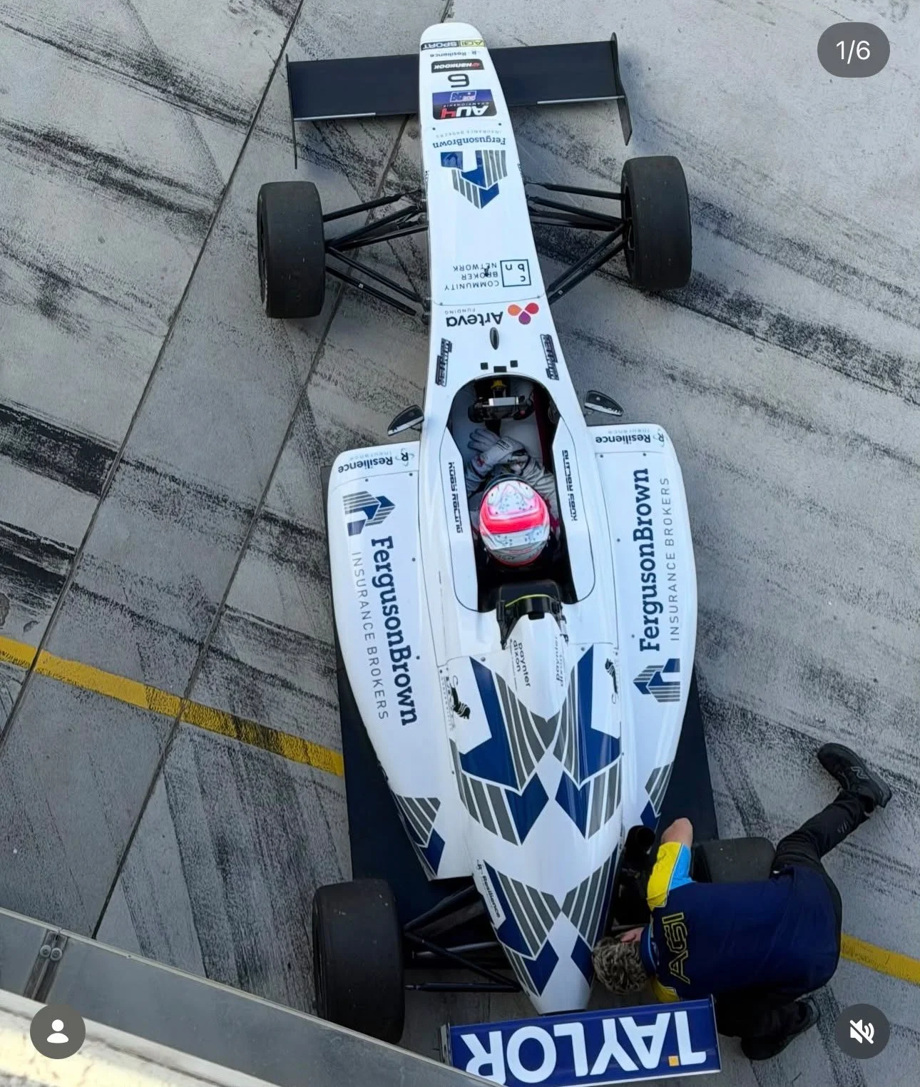 A race car with sponsorship decals, a driver in a helmet and racing suit, and team members working on or inspecting the car in a pit lane with concrete surface and yellow markings.