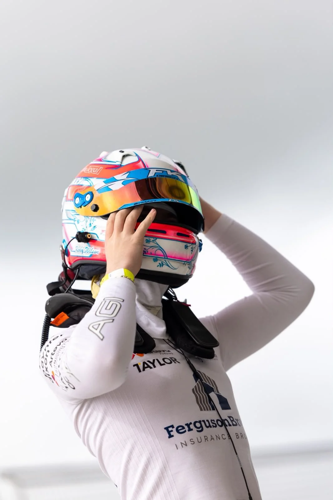 Race car driver wearing a white racing suit and colorful helmet, adjusting helmet straps.