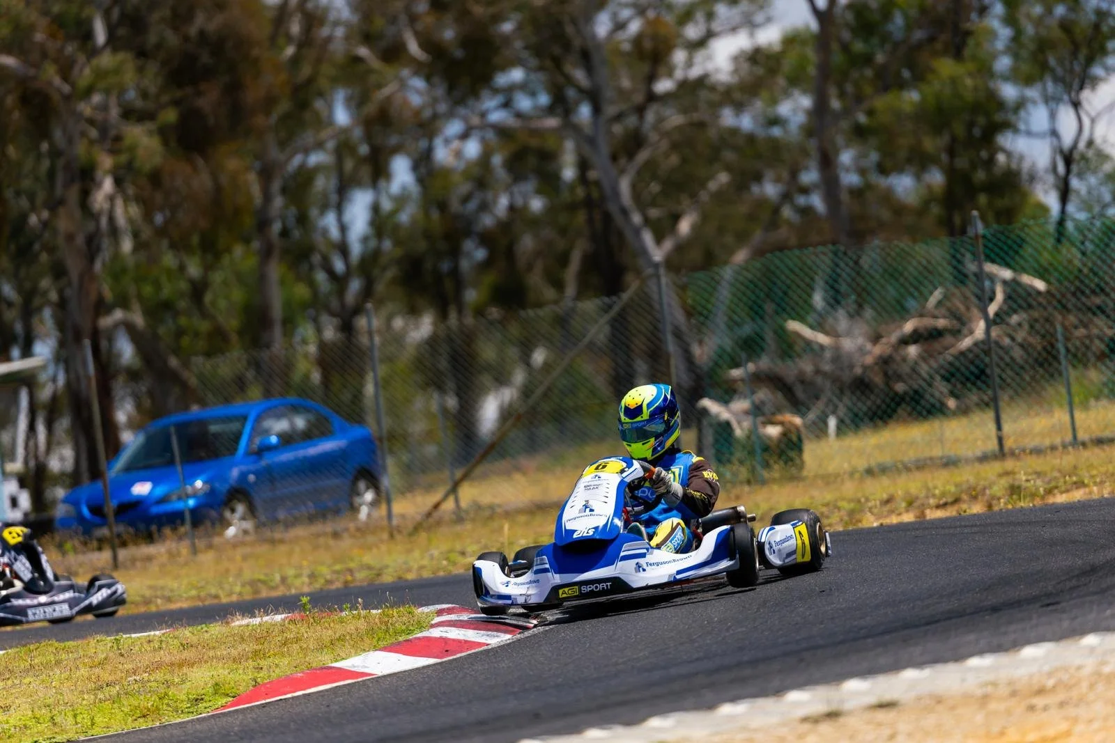 A child driving a blue and white go-kart on a race track during daytime, with another go-kart and a blue car in the background, trees, and a chain-link fence.