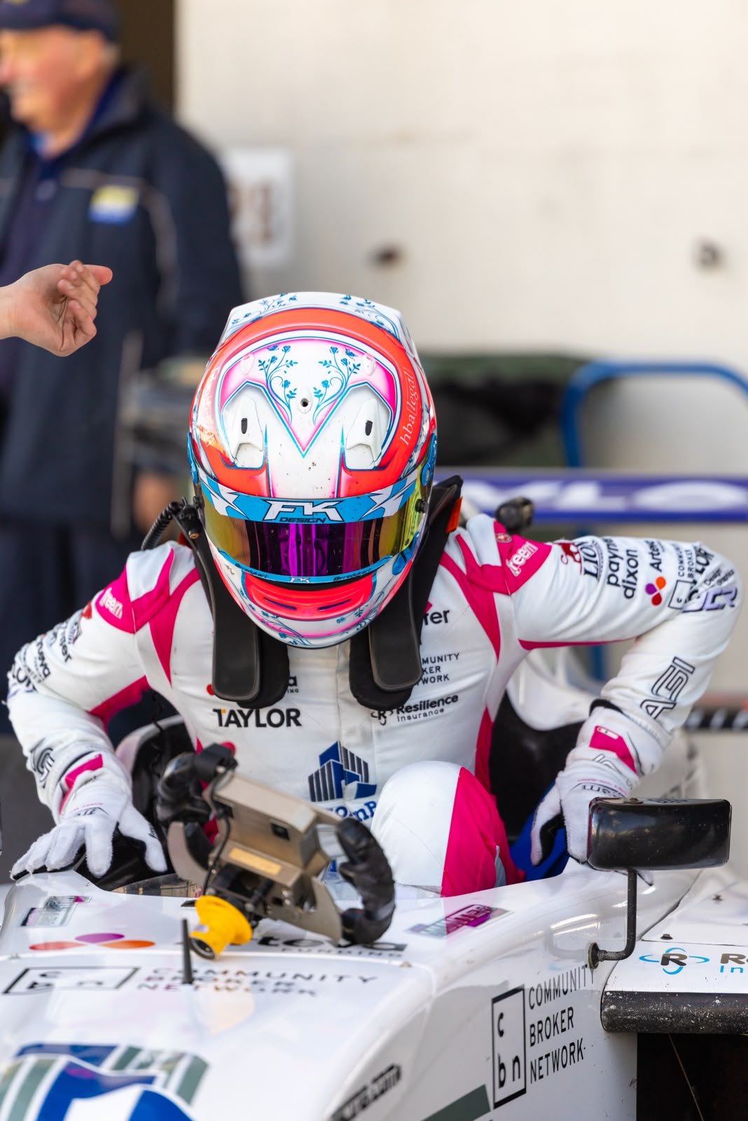 Race car driver sitting in a race car, wearing a colorful helmet and racing suit with sponsor logos, preparing for a race.