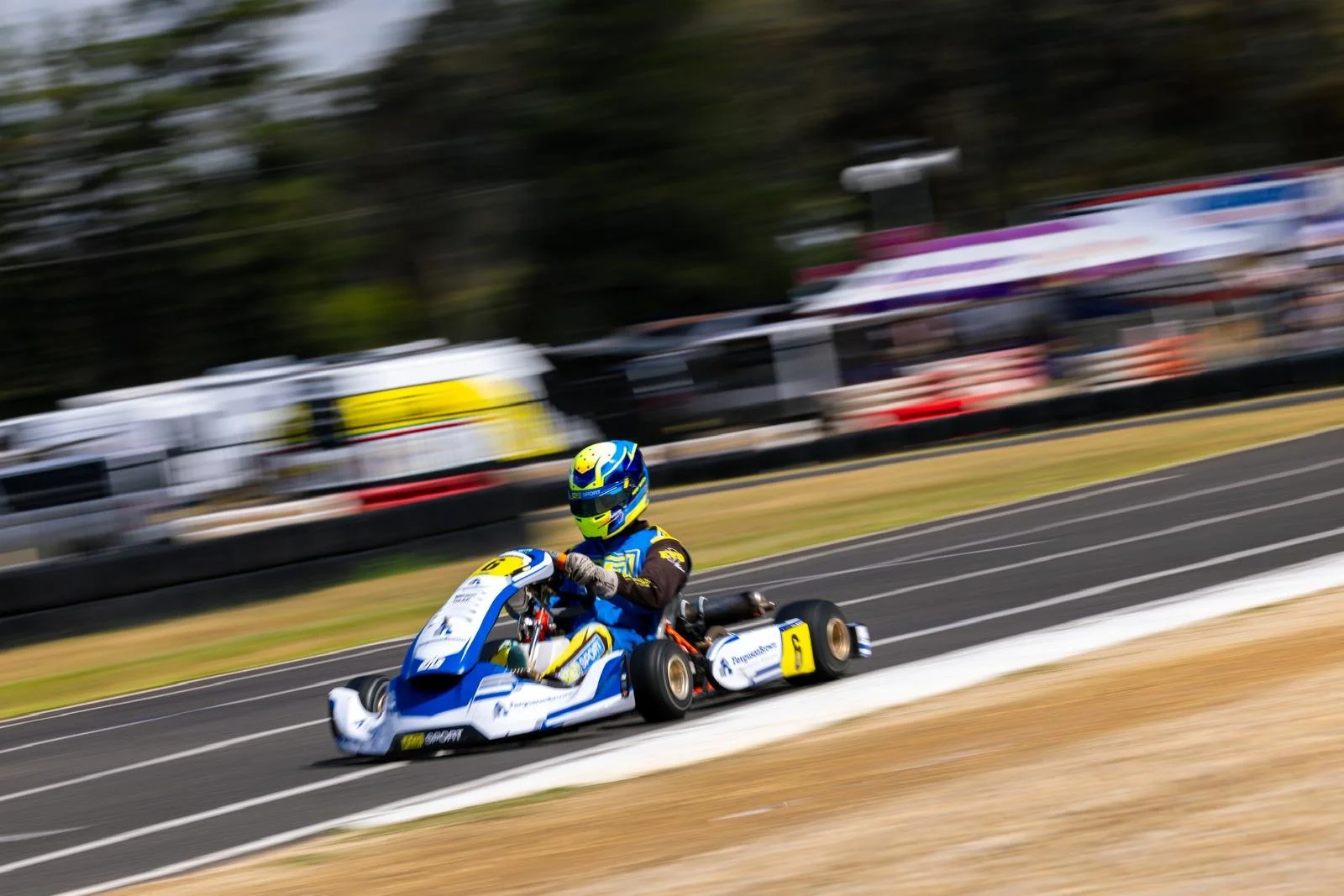 A person racing in a go-kart on a paved track with trailers and trees in the background.
