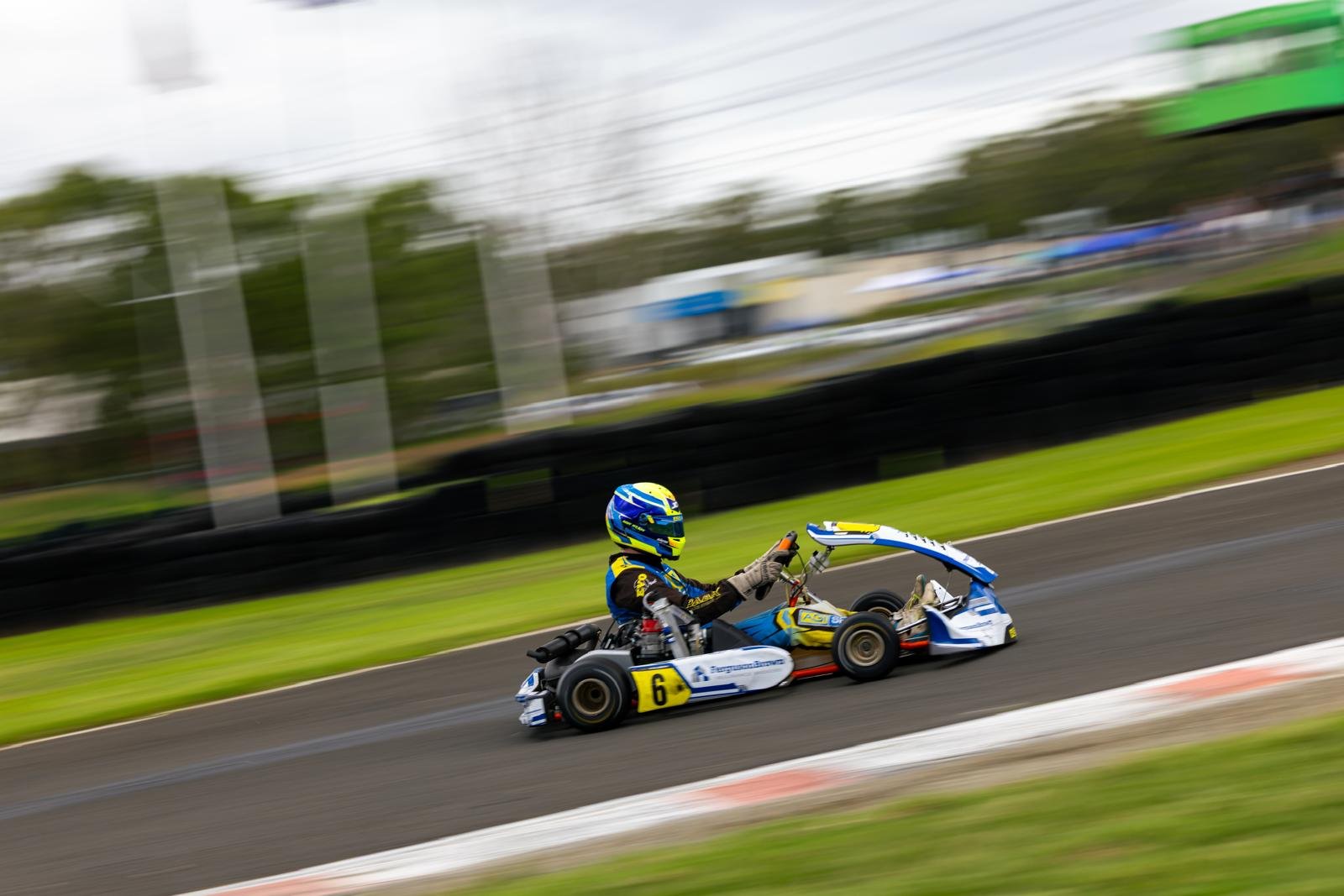 A race car driver in blue and yellow racing gear driving a go-kart on a track with motion blur background.