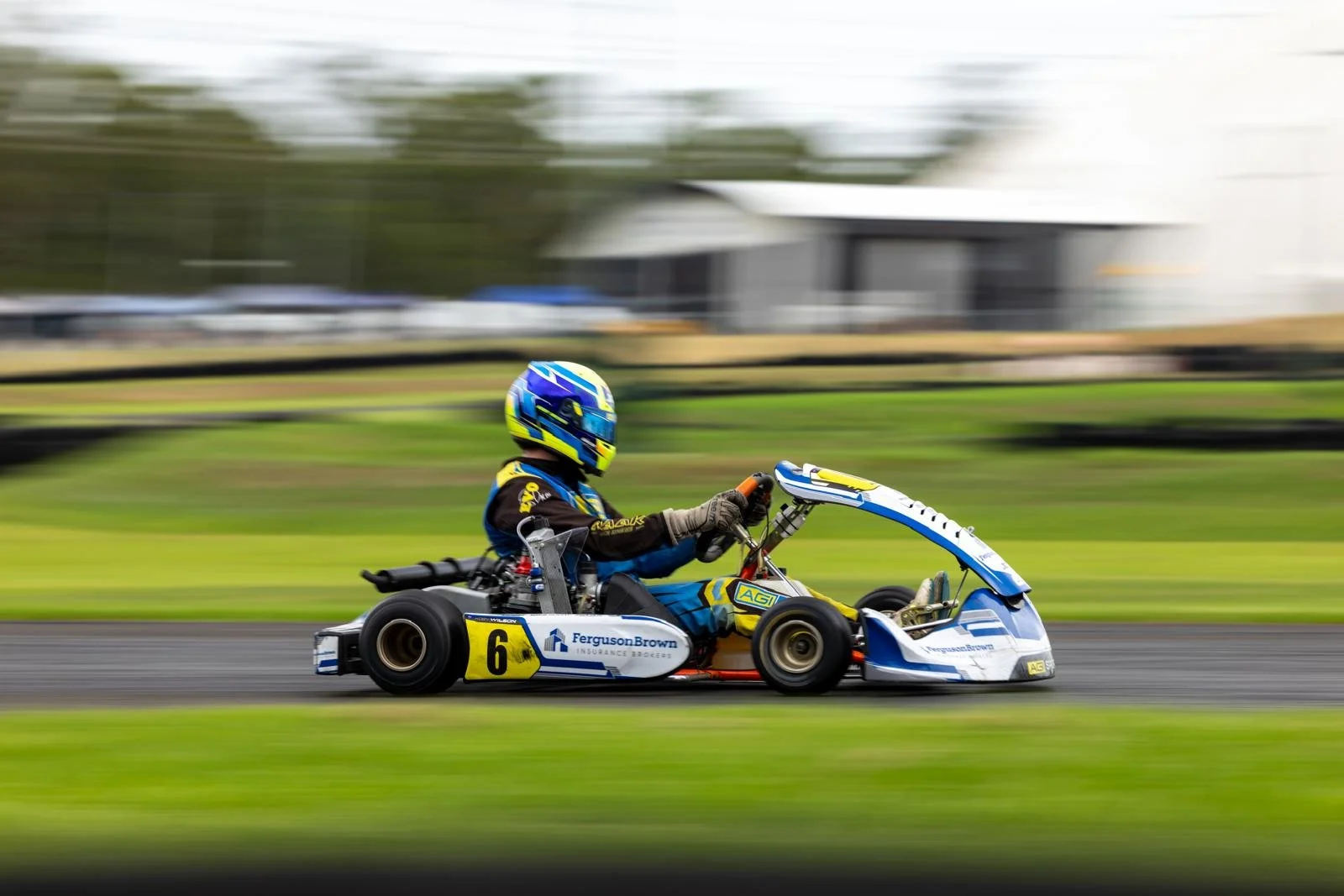 A person racing in a go-kart on a track, wearing a blue and yellow helmet and racing suit, with a blurred background indicating high speed.