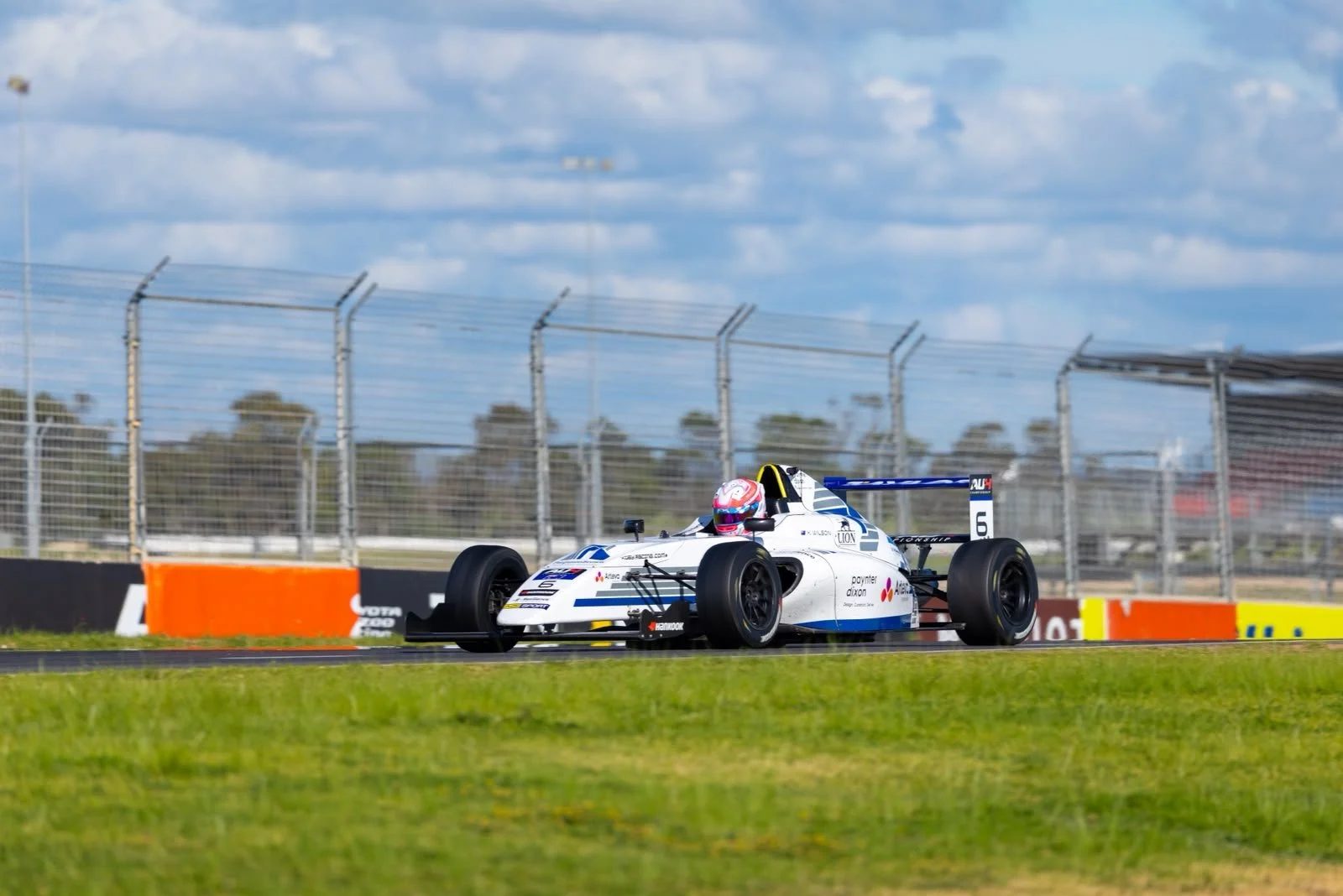 A white and blue race car on a track, with a driver wearing a pink helmet, racing near a fence and colorful barriers, under a partly cloudy sky.