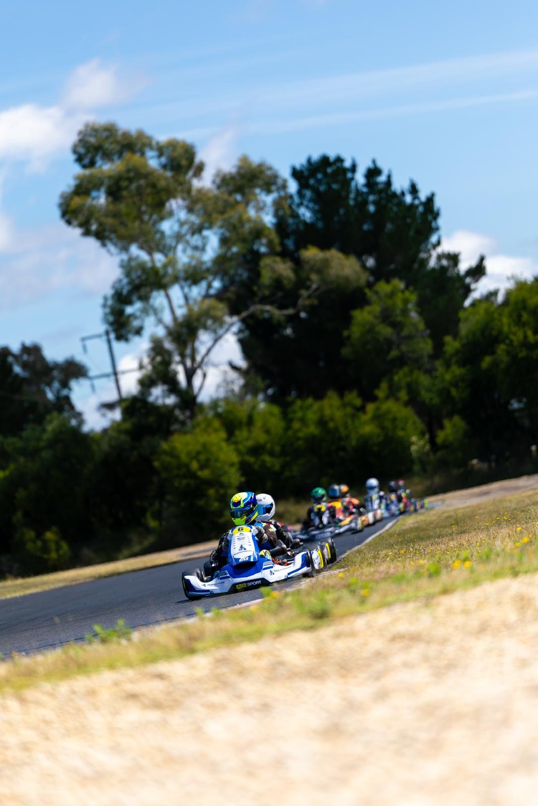 A line of go-karts racing on a track, with drivers wearing colorful helmets and suits, set against a background of trees and a blue sky.