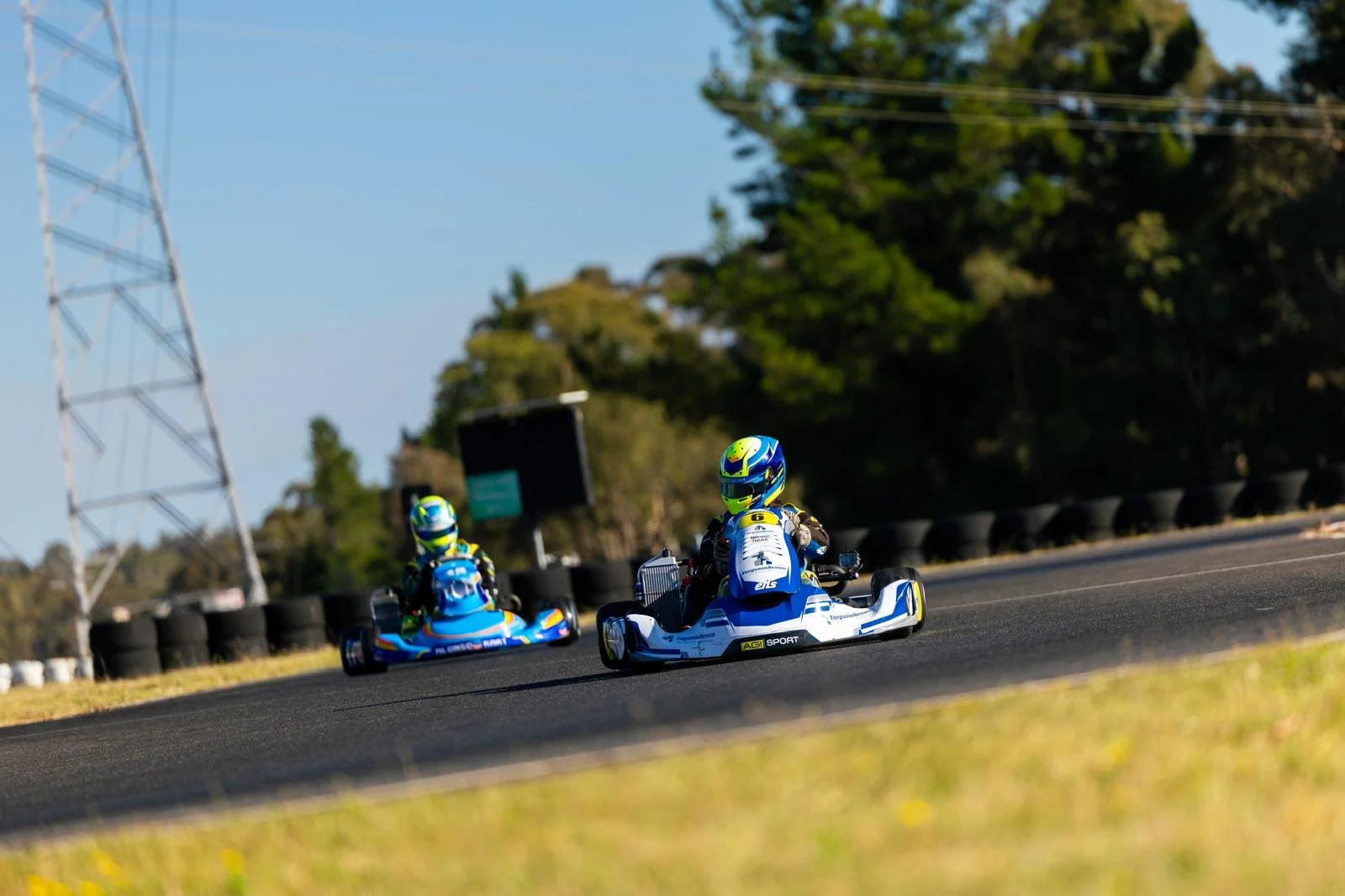 Two go-karts racing on an outdoor track, with drivers wearing colorful helmets and racing suits. The track is bordered by black tires, and there are trees and an electric tower in the background.
