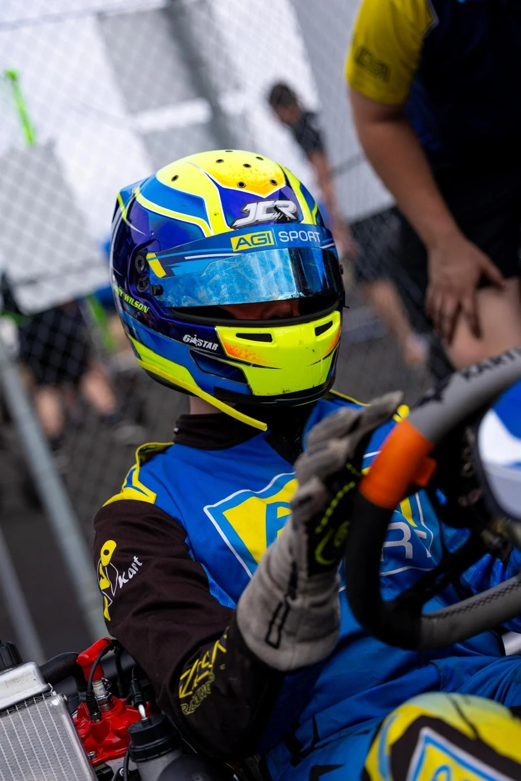 A race car driver in a yellow and blue helmet and racing suit sitting in a go-kart or race vehicle, with a blurred background of a chain-link fence and people.