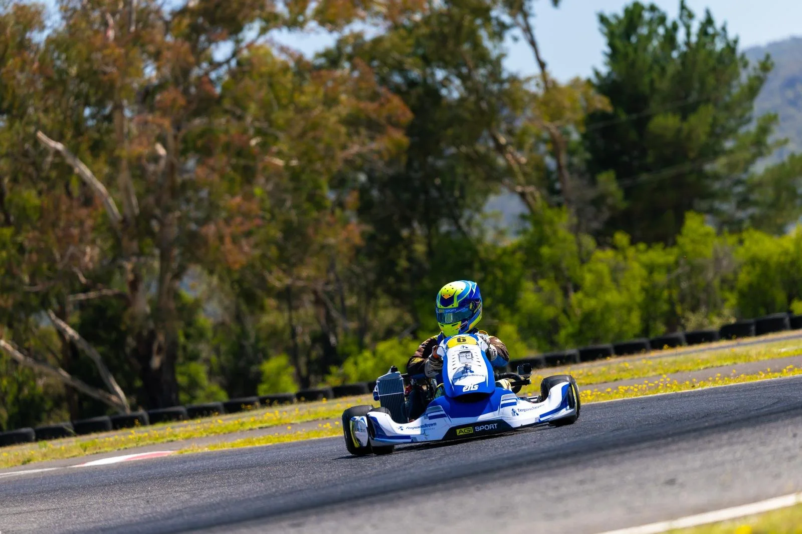 A go-kart racer on a race track surrounded by trees with autumn foliage.