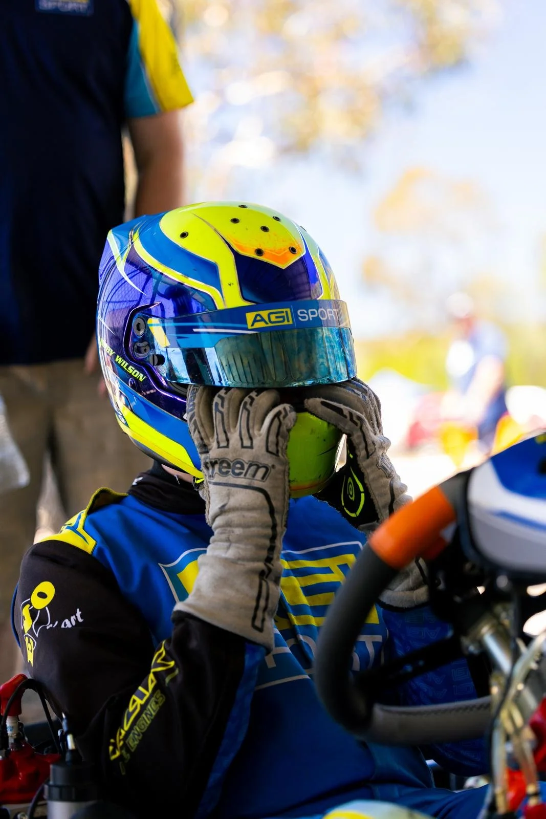 A go-kart racer in racing gear, wearing gloves and a helmet, holding a yellow ball in front of their face while sitting in a go-kart. The background shows outdoor trees and people.